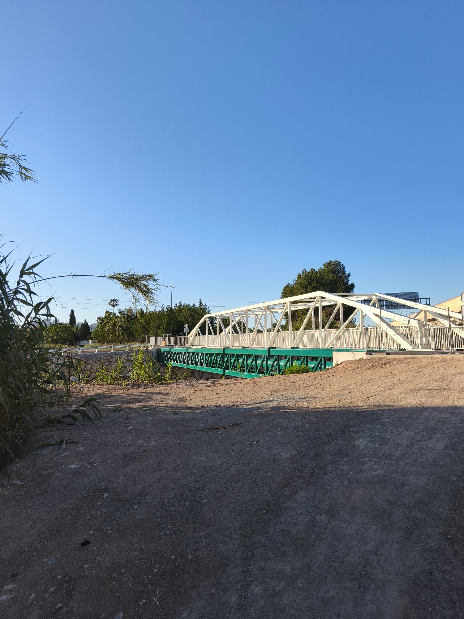 The image depicts a pedestrian bridge in Alcàsser, Spain. The bridge is white with a green undercarriage, spanning a dry riverbed. The ground is dirt and gravel. Vegetation is present in the foreground and along the riverbed. The sky is clear and blue. There are trees and buildings visible in the background. No people are visible.