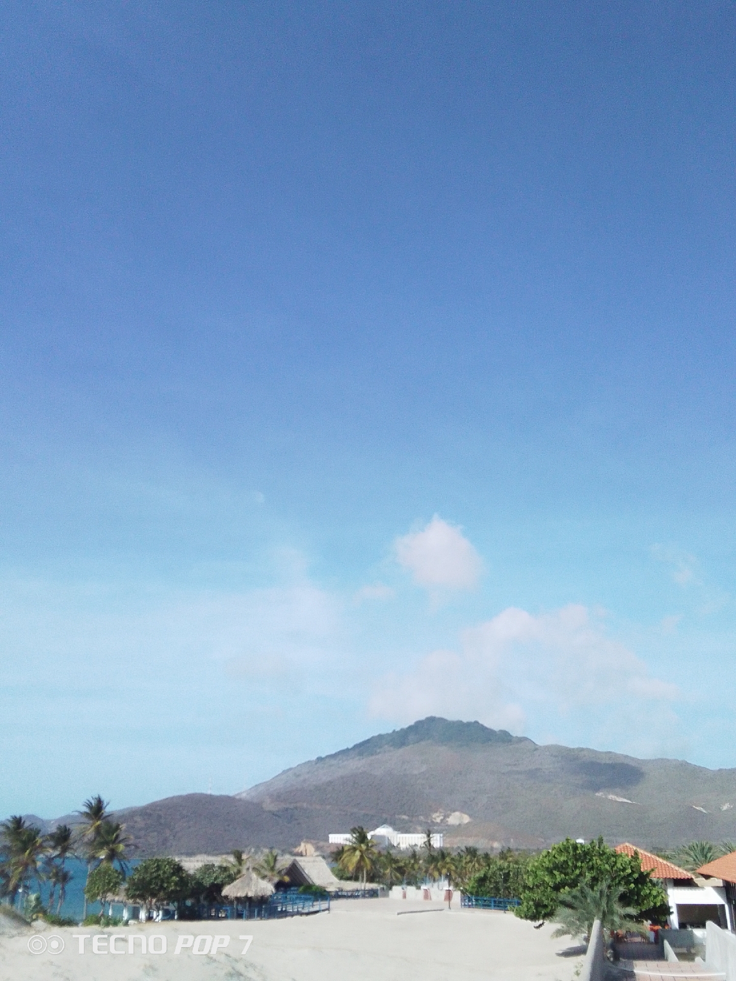 The image captures a scene in Pedro González, Venezuela, featuring a sandy foreground, palm trees, and buildings against the backdrop of a mountain. The sky is a bright blue, transitioning to a lighter hue near the horizon with scattered clouds. Several huts with thatched roofs are visible, along with a larger white building in the distance. The foreground appears to be a beach or sandy area, leading up to the trees and structures. The image has a clear, daytime setting, likely taken outdoors.