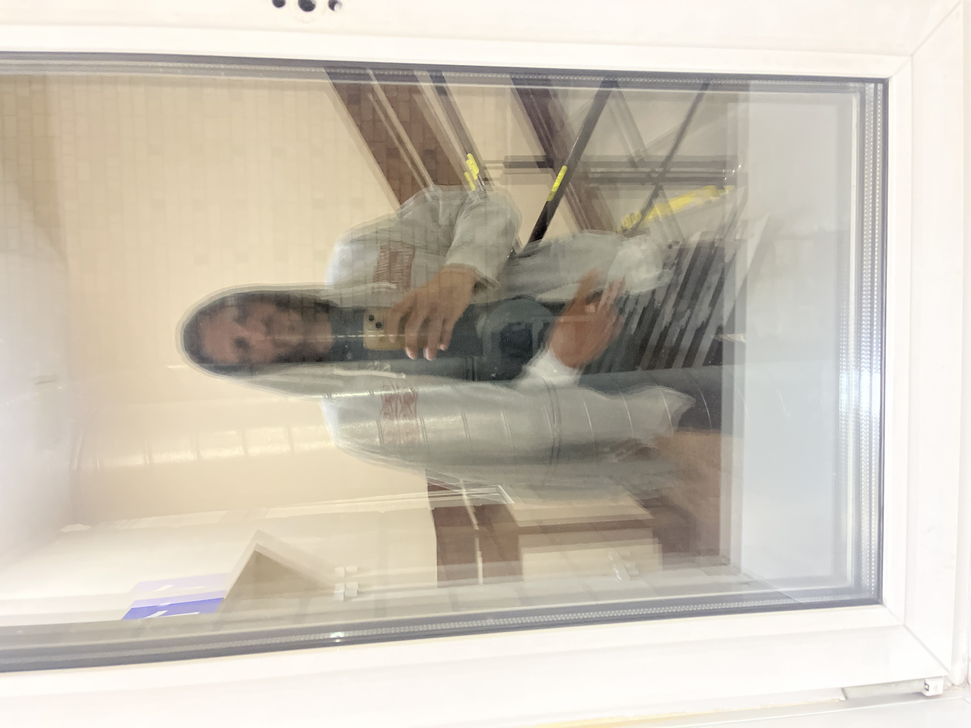 The image displays a reflection of a person taking a photograph, viewed through a clear glass panel set within a white uPVC frame. The reflection of the person dominates the central area of the frame, appearing horizontally oriented. This individual has dark hair and is wearing a light-colored, long-sleeved garment, possibly a jacket or shirt. They are holding a light-colored smartphone, likely an iPhone, in their hands, pointing it towards themselves, indicating they are capturing a selfie or a reflection shot. Their face is visible, looking towards the camera, with a neutral to slightly smiling expression.
Beyond the reflection, through the glass, an indoor environment can be seen. This background is somewhat blurred and distorted but reveals dark, angular structures, possibly support beams or ceiling components, intersecting diagonally across the upper portion of the frame. Scattered among these structures are hints of yellow objects, which could be parts of equipment or furniture. In the upper left of the reflected area, a light-toned wall with a subtle grid-like pattern is visible. Towards the bottom left, a small blue and white object, possibly a label or sign, is partially discernible. The lower right of the reflection shows a segment of what appears to be silver, corrugated flexible ductwork. The setting suggests an interior space, potentially a commercial building, office, or industrial facility. The lighting is bright and even, implying daytime. No distinct text is legible in the image.