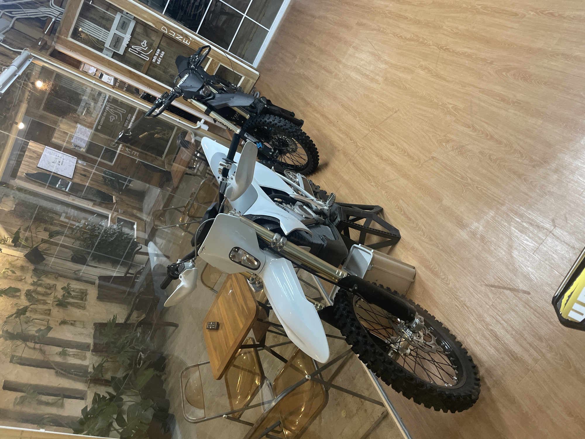 The image captures a dirt bike showroom or a similar establishment in Phu Thuy Ward, Vietnam. Two dirt bikes, one white and one black, are prominently displayed indoors. The white bike is in the foreground, angled towards the viewer, while the black bike is positioned behind and slightly to the right. The interior features a wooden floor and large glass windows that reveal a reflection of an outdoor or indoor courtyard with plants and chairs, and also glimpses of signs with Vietnamese text, suggesting the location is a commercial space, possibly a cafe or a retail store. The overall impression is one of a modern, well-lit space designed to showcase vehicles.