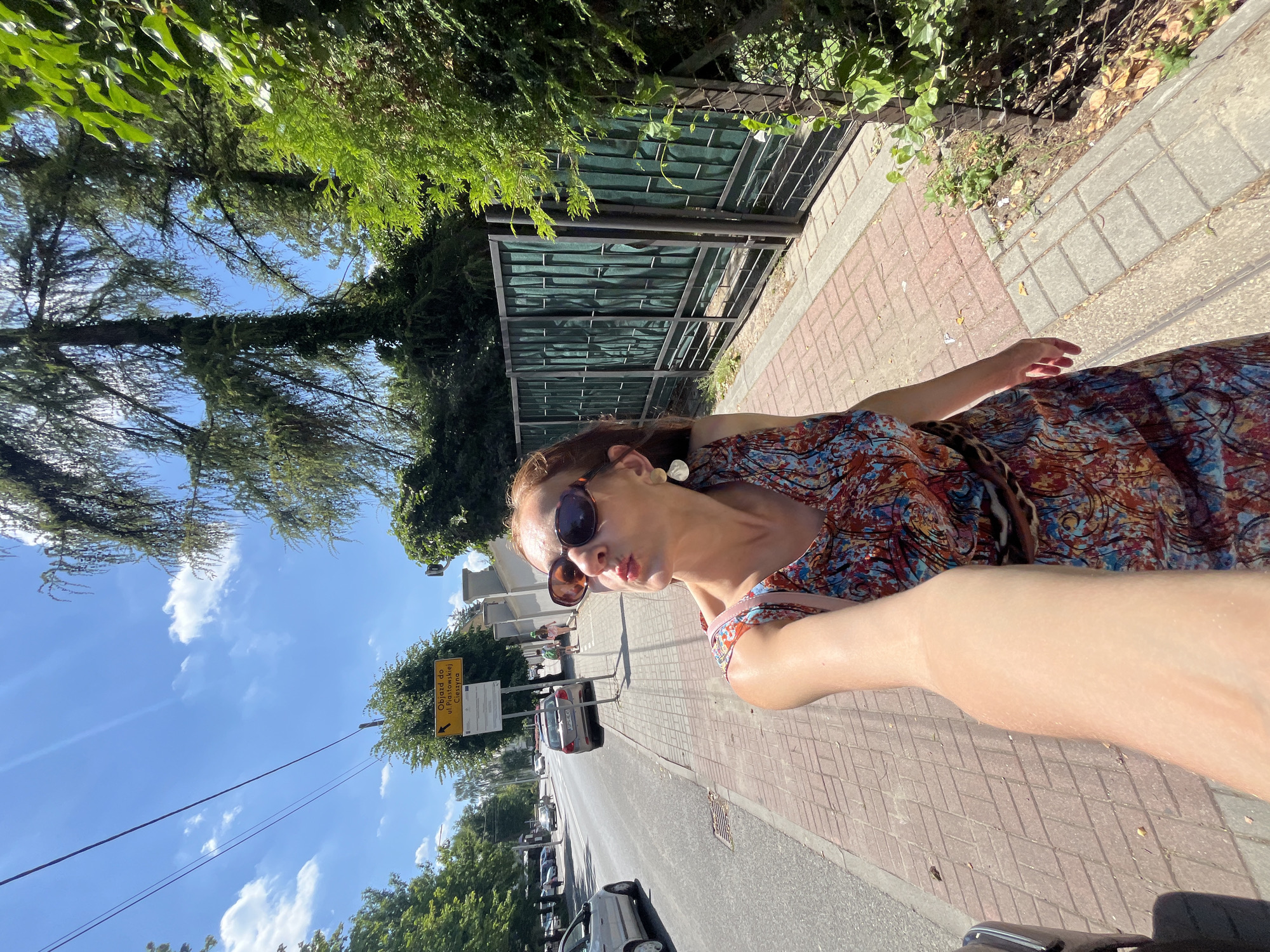 This is a vertical, candid, eye-level shot of a woman taking a selfie while standing on a brick sidewalk next to a street. She is wearing a colorful, patterned dress and sunglasses, and she is making a kissing face towards the camera. Her arm is extended, holding the camera. Behind her, there is a metal fence with green fabric coverings, trees, and a street with cars. A yellow directional sign is visible in the background. The sky is blue with some white clouds, indicating it is daytime and sunny. The scene appears to be in a town or city. The sign reads "Objazd do ul. Piastowskiej Cieszyna", which translates to "Detour to Piastowska Street, Cieszyn".