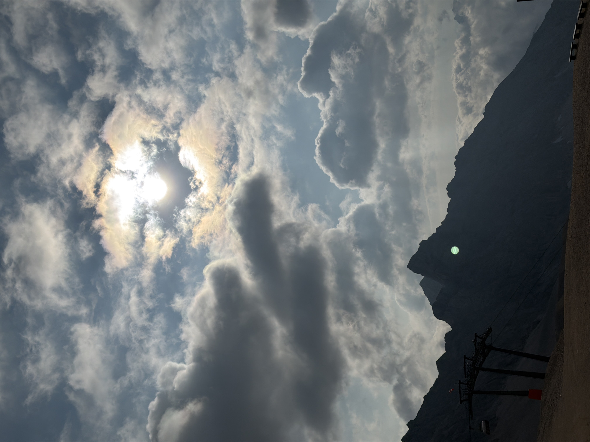 The image depicts a mountainous landscape near Garmisch-Partenkirchen, Germany, under a dramatic sky filled with clouds. In the foreground, a ski lift station with several support towers stands silhouetted against the dark mountain range. A single ski gondola hangs from a cable. The mountains themselves are dark and jagged, with the sun partially obscured by a brightly lit, iridescent cloud formation in the upper portion of the frame. A faint, circular lens flare is visible on the right side of the mountain peak. The ground in the immediate foreground appears to be a dirt or gravel path.