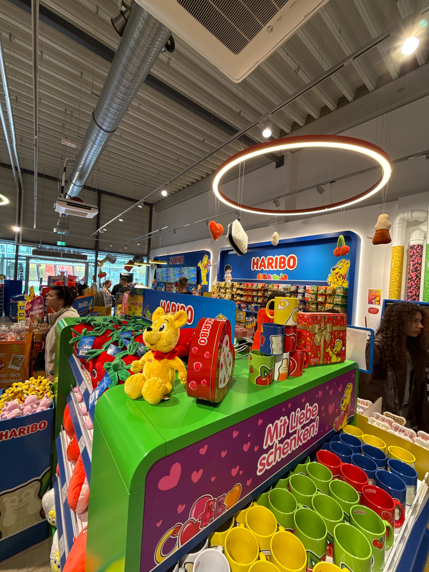 The image depicts the interior of a Haribo store in Wolfsburg, Germany. The store is brightly lit, with displays of colorful candies and merchandise. In the foreground, a display features plush toys, including a large yellow bear wearing a red bowtie, and a variety of red strawberry-shaped plush items. There are also rows of mugs in different colors, with designs related to Haribo products. Behind this, more shelves are filled with packaged Haribo candies. A blue wall with the Haribo logo and cartoon characters serves as a backdrop. A woman with curly brown hair is visible on the right side of the image, looking to her left. Other customers and staff are faintly visible in the background. The ceiling shows industrial ventilation ducts and a circular light fixture. The overall scene is characteristic of a retail environment selling confectionery and branded merchandise.