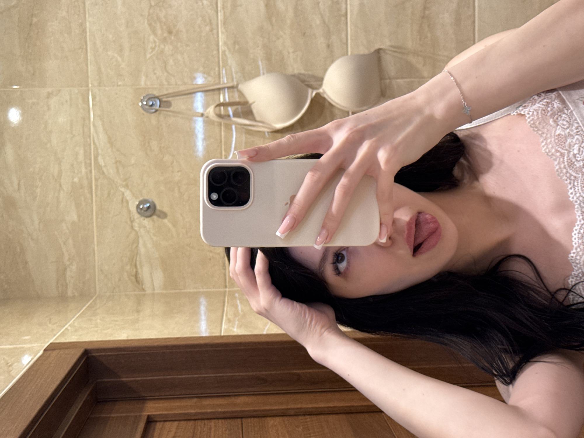 A young woman with dark hair is taking a selfie in a bathroom with beige tiled walls. She is holding a smartphone in her right hand, obscuring the left side of her face. Her left hand is raised to her head. Her tongue is sticking out, and her eyes are looking upwards. A beige bra is hanging on the wall behind her. A wooden door is visible to the left of the frame.