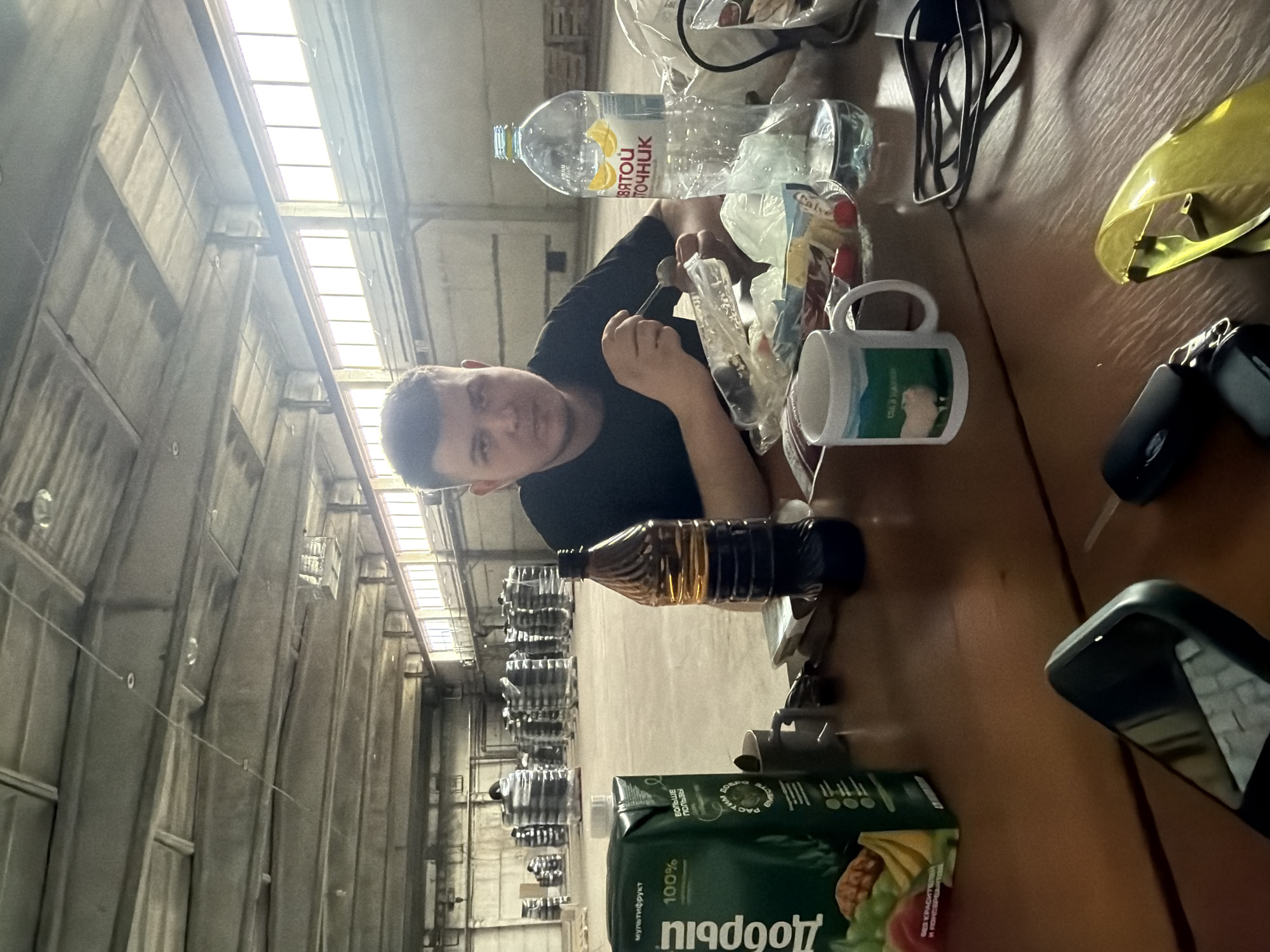 A man is seated at a table in a large industrial building with high ceilings and many rows of stacked cylindrical objects in the background. The table has various items on it: a bottle of water, some food, a mug, a bottle of what appears to be oil, a carton of juice, sunglasses, keys, and a cell phone. The man is looking at the camera. The OCR text in the image indicates that the water bottle is labeled "ВЯТОЙ ТОЧНИК" and the juice carton is labeled "Добрый". The juice carton also contains the words "мультифрукт" and "100%". There is nothing in the image that indicates the image was taken in the city of Шебунино, Russia.