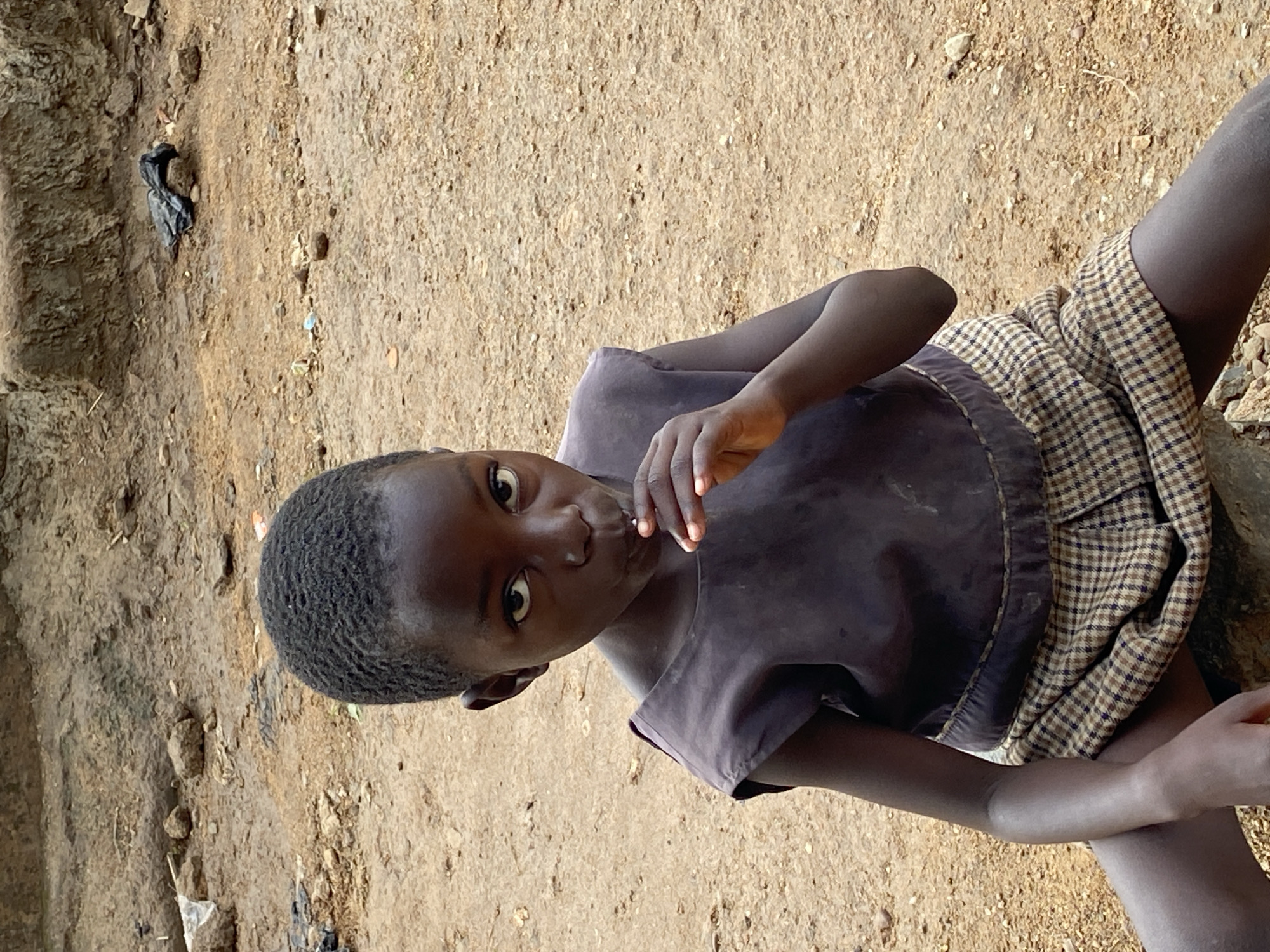 This image is a direct portrait of a young child, captured outdoors in what appears to be a rural or undeveloped area in Riyom, Nigeria. The child, with dark skin and short, dark, closely-cropped hair, is positioned prominently in the frame, looking directly at the camera with wide, attentive eyes and a neutral, slightly curious expression.
The child is wearing a dark purple or grey short-sleeved top and a brown and white checkered skirt or wrap-around garment. One hand is raised towards their mouth, with fingers gently touching their lips, possibly holding a small, unidentifiable item or simply resting in a thoughtful pose.
The background and ground consist primarily of dry, light brown earth or dirt, scattered with small rocks, pebbles, and some minimal debris, including a crumpled dark plastic item visible on the left. The scene suggests a dusty, unpaved environment. The lighting is bright and even, indicative of daytime, without harsh shadows, implying either an overcast day or a time when the sun is not directly overhead. There is no visible text in the image. The overall impression is a candid moment, highlighting the child's presence within their natural surroundings.