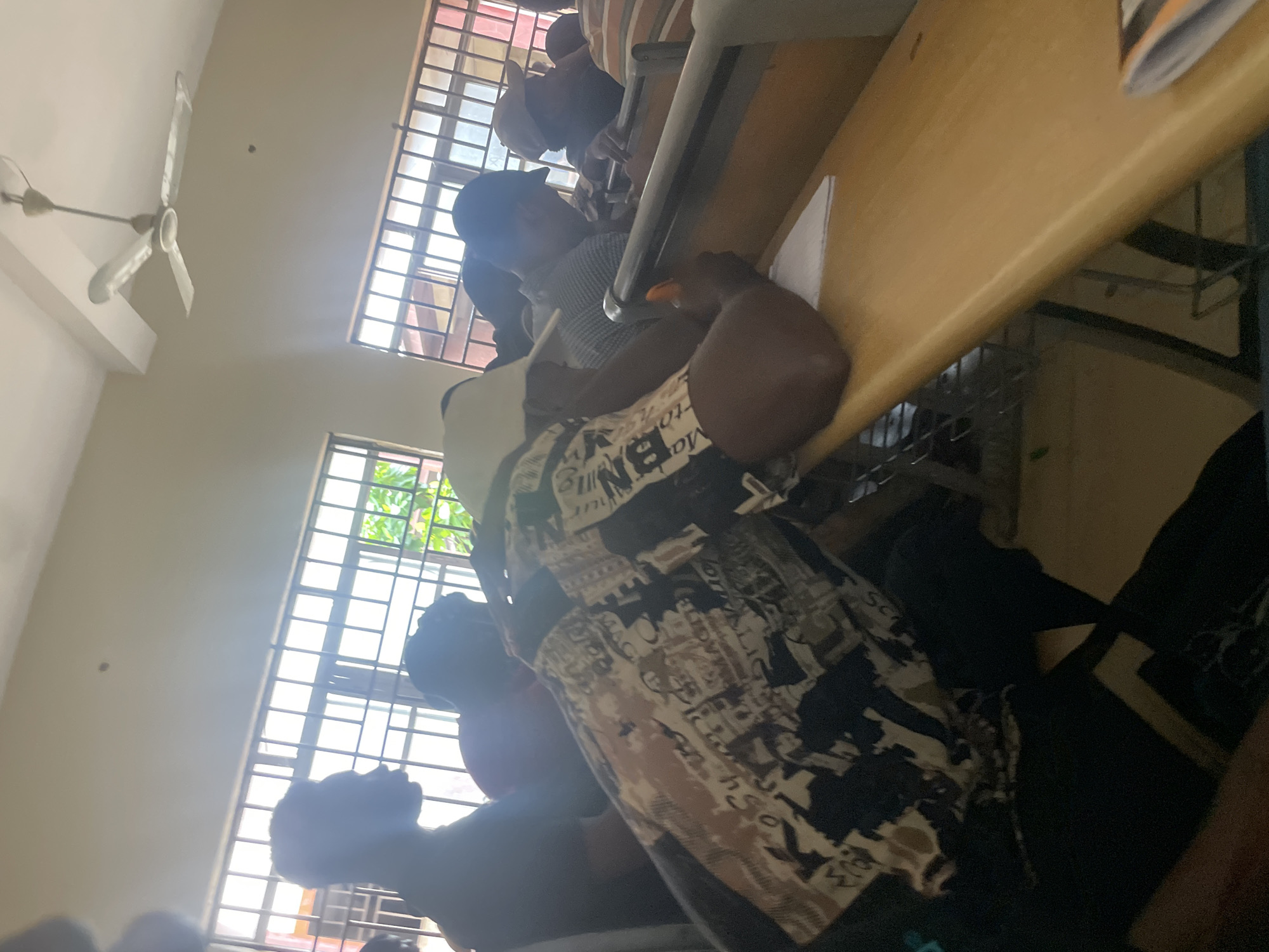 The image captures an indoor scene, possibly a classroom, filled with people seated at desks. The overall lighting suggests an ambient indoor setting, with light coming from windows that appear to have bars. In the foreground, a person wearing a patterned shirt and a white cap is leaning on a wooden desk, holding a pen. Other individuals are visible in the background, some wearing hats or caps, and appear to be engaged in activities at their desks. A ceiling fan is visible in the upper portion of the image. The presence of desks and the number of people suggest an educational or meeting environment. The details of the location in Ihiagwa, Nigeria, cannot be determined solely from the visual information presented in the image.