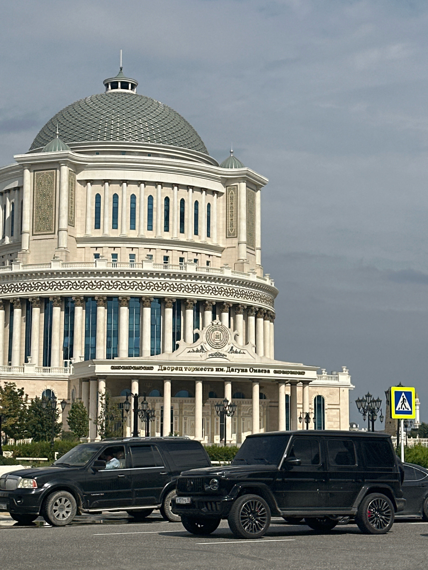 In the city of Grozny, Russia, on a cloudy day, a large, ornate building with a prominent dome stands in the background. The building's facade features classical architectural elements, including columns, arched windows, and decorative stonework. Below the main structure, text is visible on a frieze, reading "Дворец торжеств им. Дагуна Омаева" (Palace of Celebrations named after Dagun Omaev).
In the foreground, two black SUVs are parked on a paved road, suggesting a street scene. The SUV on the left is a Lincoln Navigator, and the one on the right appears to be a Mercedes-Benz G-Wagon. A pedestrian crossing sign with a yellow background and a blue symbol of a person walking is visible to the right, indicating a crosswalk. The overall scene suggests a public area in Grozny, possibly near a significant landmark or venue.