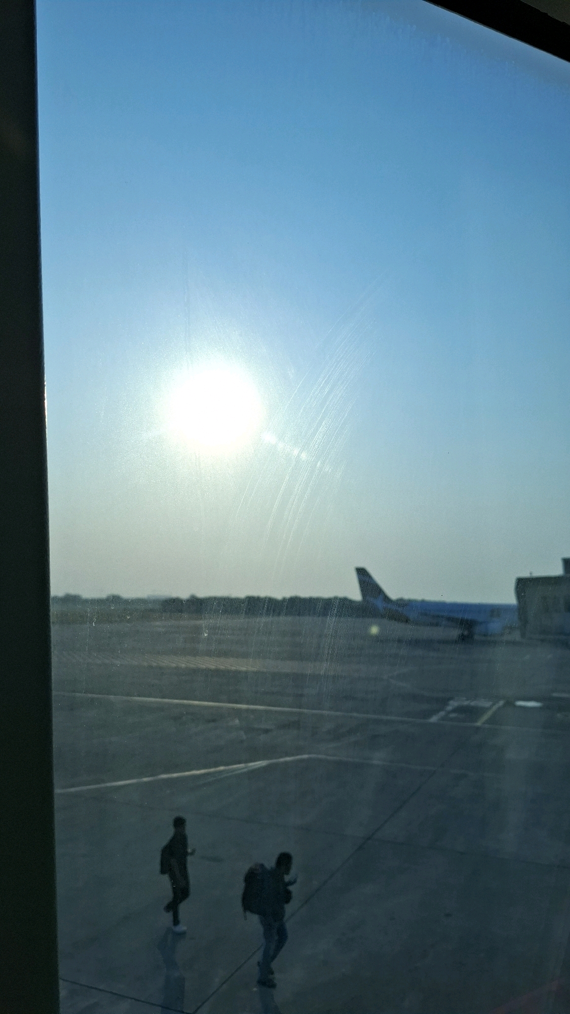 The image depicts an outdoor scene, likely an airport tarmac, under a bright, hazy sky. A large, luminous sun is prominent in the upper left quadrant of the image, casting a glare and lens flare that obscures some details. In the mid-ground, a commercial airplane is visible, parked or taxiing on the tarmac. The plane is light-colored with a dark tail fin. A building or structure, appearing as a light-colored rectangular block, is situated to the right of the airplane. The tarmac itself is dark and appears wet, reflecting the sky and the sun. In the foreground, two individuals are walking across the tarmac, both carrying backpacks. The person on the left is walking in a more upright posture, while the person on the right is leaning forward slightly. Their reflections are visible on the wet surface. The overall impression is that of a travel or departure scene, possibly at dawn or dusk due to the sun's low angle and the hazy atmosphere. The scene is viewed through a window, as evidenced by the dark vertical frame on the left edge of the image and faint horizontal lines across the glass. The location is stated to be the city of Semarang, Indonesia, suggesting this is likely the Ahmad Yani International Airport (SRG).
