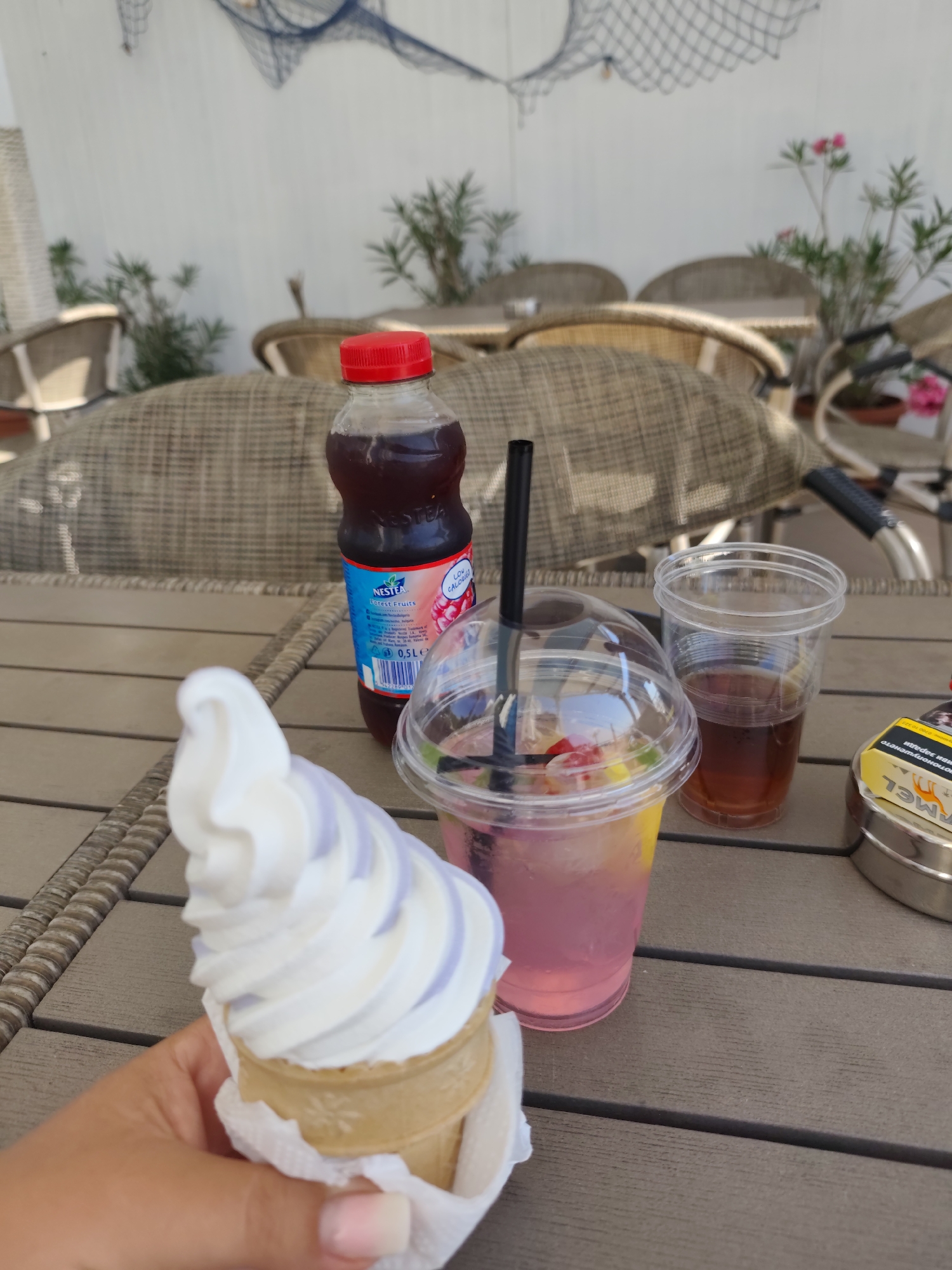 The image shows a close-up view of a table with several items on it, set against a blurred background of outdoor seating. In the foreground, a hand is holding an ice cream cone with white and light purple swirls of soft serve. To the right of the ice cream, there is a clear plastic cup filled with a pink beverage containing fruit and ice, with a black straw. Next to that, another clear plastic cup contains a dark amber liquid, likely iced tea or soda. A bottle of Nestea "Forest Fruits" beverage with a red cap stands behind the pink drink. On the far right, a portion of a silver ashtray containing a yellow packet of Camel cigarettes is visible. The table appears to be made of wood planks. The background suggests an outdoor seating area with woven chairs and tables, and a white wall with decorative fishing nets and plants. The overall impression is a relaxed outdoor refreshment setting, possibly at a cafe or restaurant in Nesebar, Bulgaria, given the context provided.