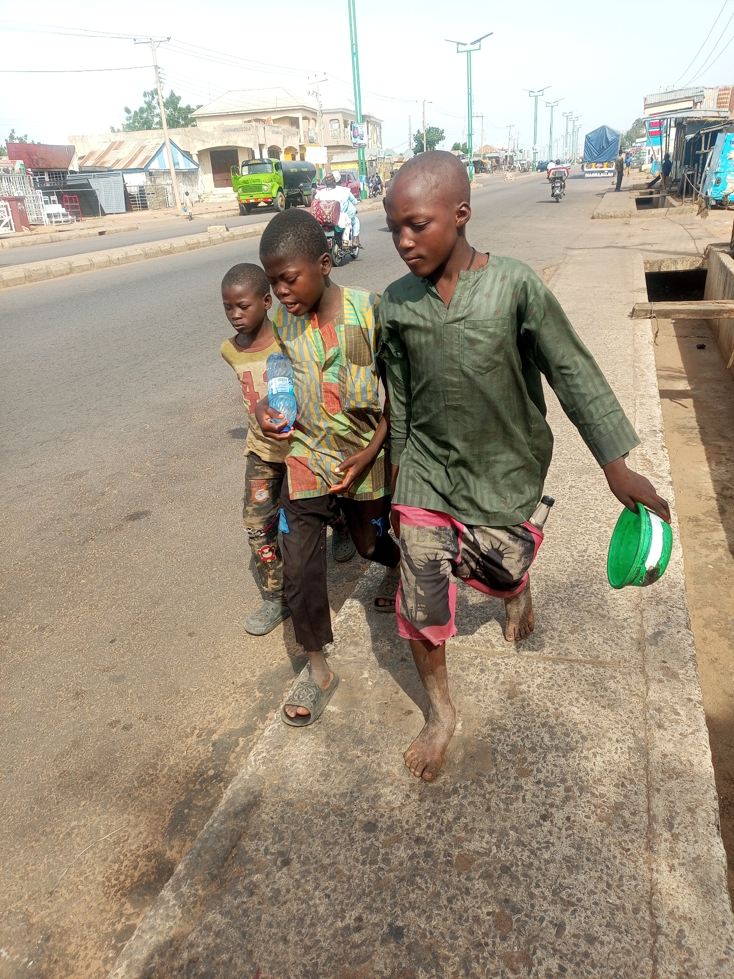 Three young boys are walking on a concrete sidewalk next to a paved road in Garko, Nigeria. The boy on the right, wearing a green long-sleeved shirt and patterned shorts, is carrying a green bowl in his right hand. He is barefoot. The boy in the middle, wearing a multicolored patterned shirt and dark pants, is holding a clear plastic water bottle. He is wearing grey sandals. The boy on the left, wearing a yellow and white striped shirt and camouflage pants, is also barefoot. The road is busy with traffic, including a green truck and several motorcycles. Buildings line the opposite side of the road, and a utility pole with streetlights stands near the sidewalk. The sky is bright and appears to be daytime.
