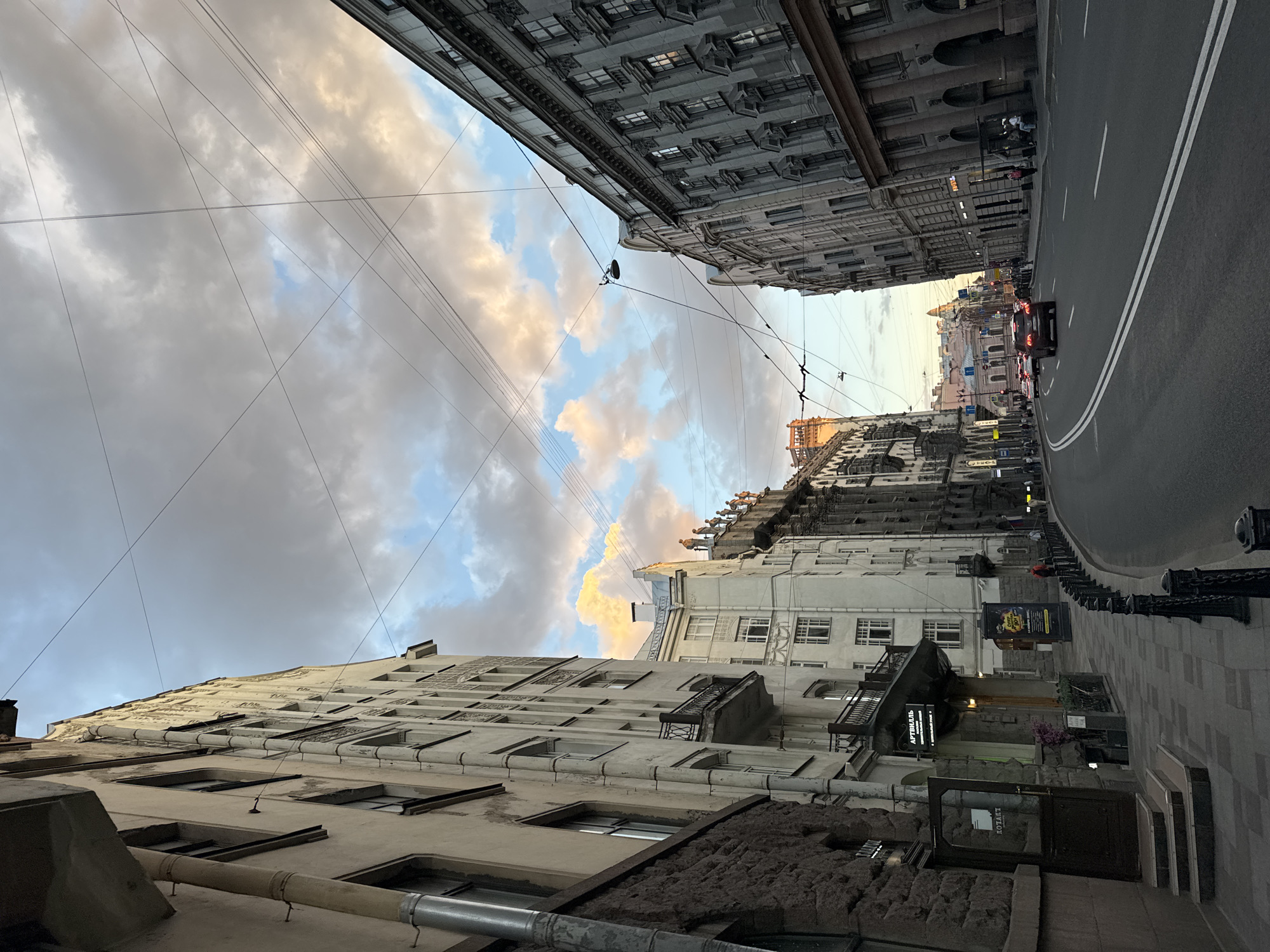 The image depicts a street scene in Saint Petersburg, Russia, characterized by its neoclassical architecture and a sky filled with dramatic clouds tinged with sunset colors. The perspective is from the sidewalk looking down a wide, paved street lined with multi-story buildings. On the left, a building with a stone-clad lower level and a lighter facade above features several windows and a prominent drainpipe. A dark doorway with a glass panel leads into what appears to be a business with signage. Further down the sidewalk, there are decorative iron fences and a few people can be seen walking.
The street itself is asphalt with double white lane markings, indicating a two-way traffic flow. Several cars are visible on the road, including a dark red SUV in the foreground. The buildings on both sides of the street are imposing and ornate, with numerous windows, balconies, and architectural details typical of European city centers. The building on the right is particularly striking with its colonnaded ground floor and repetitive window patterns on upper levels.
The sky is a prominent feature, with a mix of dark, heavy clouds and patches of blue sky illuminated by the setting sun, creating a dynamic and somewhat moody atmosphere. Numerous overhead wires crisscross the sky, suggesting an urban infrastructure with power or communication lines. In the distance, the street curves and disappears, with hints of more buildings and possibly traffic lights. The overall impression is of a historic and bustling urban environment during the twilight hours.