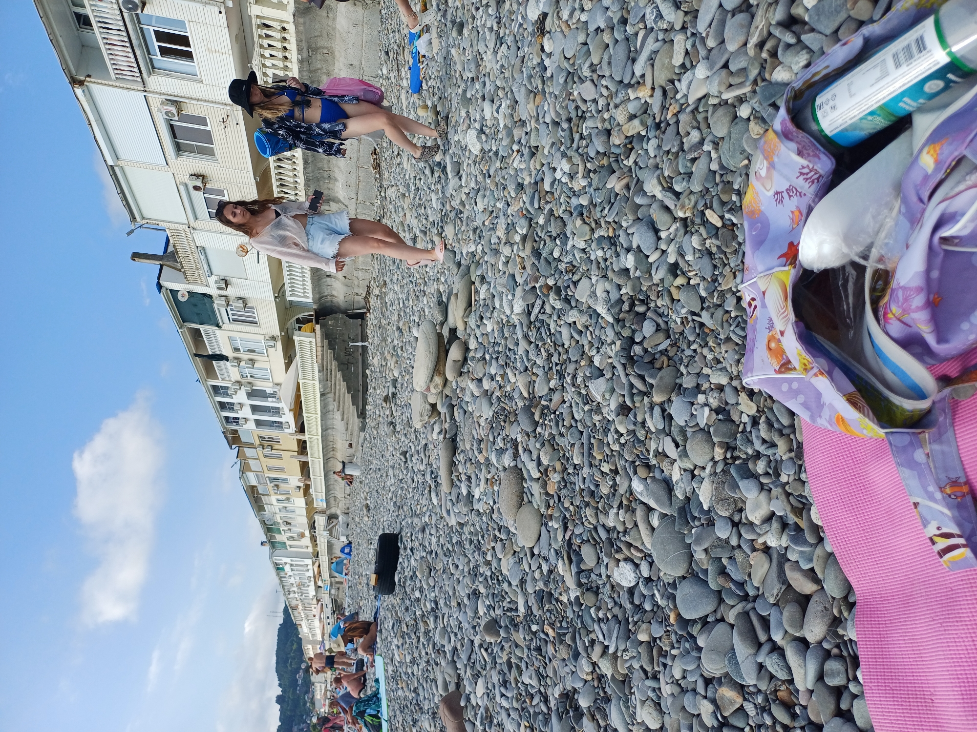 The image depicts a bustling pebble beach in Sochi, Russia, with the Black Sea and a row of hotels in the background. The foreground features a pink yoga mat and a partially visible bag filled with beach essentials, including a bottle and other items. In the middle ground, two young women are walking along the shore. The woman on the left wears a light, sheer shirt and denim shorts, while the woman on the right is dressed in a blue bikini and a patterned jacket. Numerous beachgoers are scattered across the pebbles, some sunbathing, others conversing, and some seen in the background under small tents. The sky is partly cloudy, indicating a pleasant day at the beach.