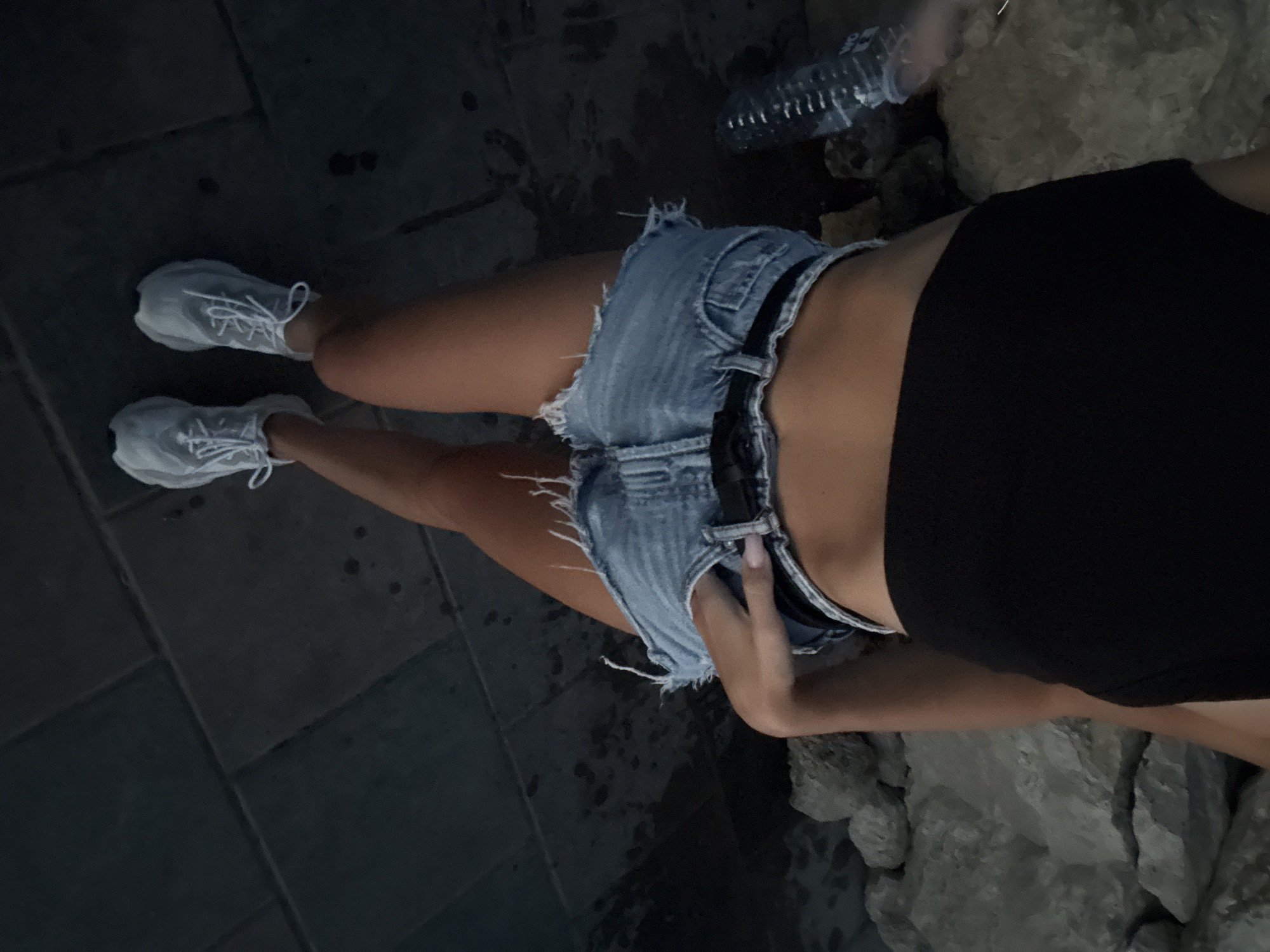 A person stands on a dark stone pathway, their legs and torso visible from the mid-torso down. They are wearing ripped denim shorts, a black crop top, and light grey sneakers with white laces. A hand is tucked into the front pocket of the shorts. The background consists of large, light-colored rocks on the left and right sides, with a partially visible plastic water bottle on the far left. The lighting suggests it is dusk or evening. The scene is indoors or outdoors, as indicated by the pathway and rocks. The specific location in Cimișlia, Moldova cannot be determined from the visual information provided.