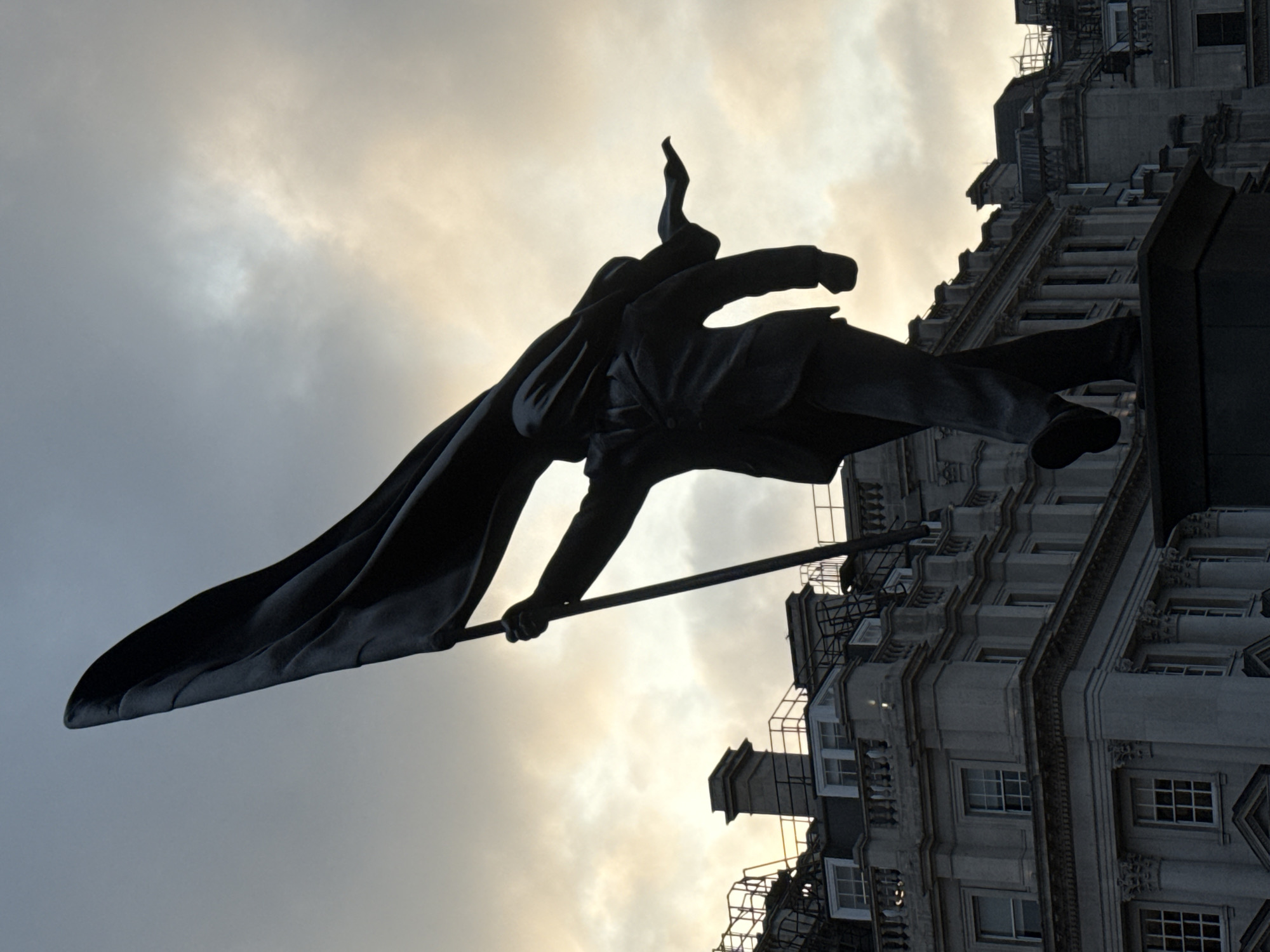 A low-angle, rotated shot focuses on the statue atop the Cavell Van in St Martin's Place, London, against a backdrop of sky and a building facade. The statue, silhouetted against the sky, depicts a figure holding a flag-like object. The building behind has a multi-story facade with windows and architectural details. The sky shows a mix of clouds and light, suggesting either sunrise or sunset. The entire scene has a subdued color palette due to the backlighting.
