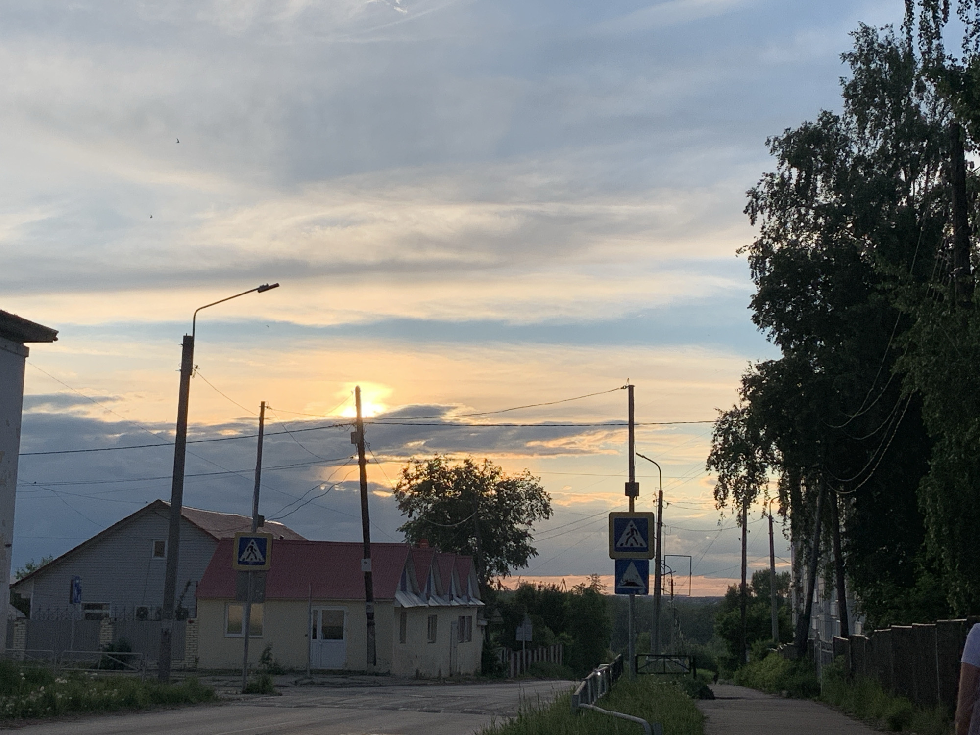 The image depicts an urban scene in Chaikovsky, Russia, likely a residential area at dusk or dawn. The sky is filled with clouds, transitioning from blue to orange and yellow near the horizon, where the sun is obscured. A two-lane asphalt road curves slightly to the right in the foreground.
To the left of the road is a light-colored building with a red roof. A pedestrian crossing sign is posted near the building. Utility poles run along the road, carrying power lines. On the right side of the road is a sidewalk partially obscured by overgrown vegetation. A metal guardrail runs along the edge of the sidewalk. Tall trees with dense foliage line the right side of the image.
In the distance, beyond the trees, there is a glimpse of more urban landscape. A person wearing a white shirt is partially visible on the extreme right edge of the frame. The scene suggests a quiet, possibly suburban environment within the city.