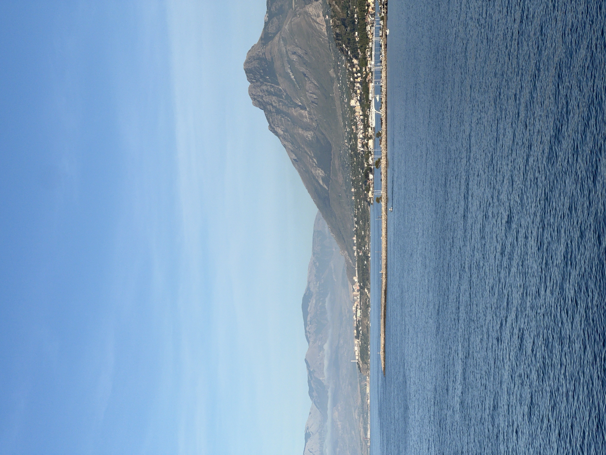 This vertical image depicts a panoramic coastal scene under a clear blue sky, identified as being located in or near the city of Sperone, Italy.
The lower half of the image is dominated by an expanse of deep blue, slightly rippled water, indicating a calm sea surface. Extending from the right mid-ground into the water is a long, curved stone breakwater, appearing light grey. A small, light-colored boat with a single occupant is visible on the water to the left of the breakwater's end. Another small, white boat is visible closer to the marina on the right.
Above the breakwater, the coastline reveals a densely developed urban area, forming a town or city. Numerous buildings, predominantly light-colored (white, cream, pale grey) with some red-tiled roofs, extend along the shore and up the lower slopes of the adjacent mountains. A prominent marina is visible behind the breakwater, crowded with numerous moored sailboats and yachts, identifiable by their white masts and structures. An elevated white structure, possibly a bridge or walkway, spans part of the marina. Towards the left of the town, a tall, slender, white industrial-style stack or chimney rises above the buildings.
The background is characterized by a range of mountains. A large, prominent, rugged mountain with dark grey and greenish-brown hues dominates the right side of the frame, showcasing visible rock formations and slopes. To the left, a series of more distant, lighter grey-blue mountains extends into the haze, with low-lying clouds or mist obscuring their bases. The sky above is a gradient of clear blue, transitioning from a slightly deeper blue at the top to a lighter hue near the horizon, with minimal atmospheric haze. No other discernible people, specific actions, or significant interactions are evident.