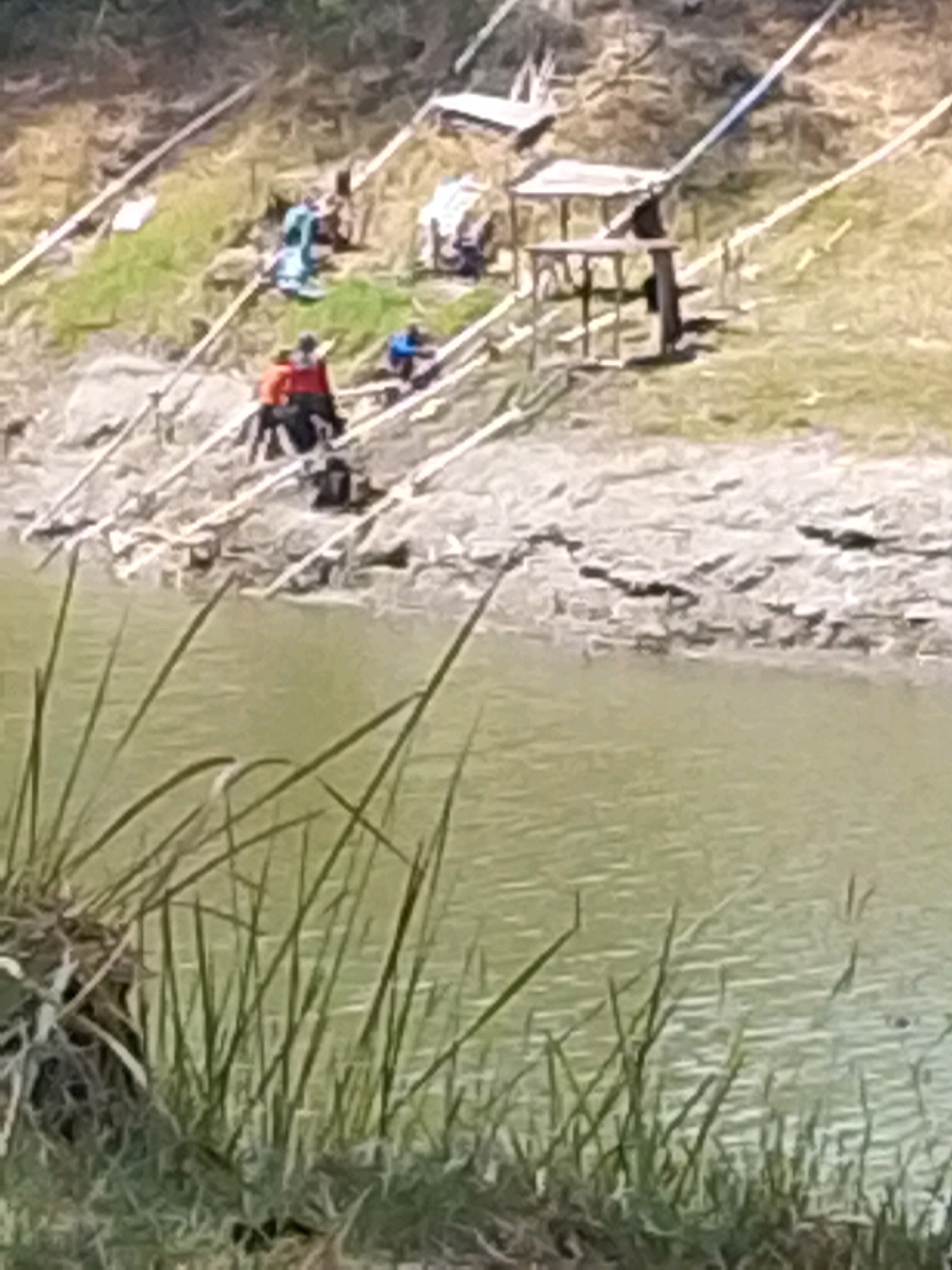 The image depicts a riverside scene in Grebegan, Indonesia, with several individuals engaged in what appears to be fishing or recreational activities. The foreground is dominated by tall grass and the muddy bank of a body of water, likely a river or lake, with calm, greenish water. In the midground, on a sloped, grassy embankment, a group of people are visible. Some are seated, and others are standing near wooden structures that resemble makeshift fishing platforms or shelters. One individual, wearing a red top and dark bottoms, is clearly visible. Other figures, dressed in blue and white, are also present, some appearing to be casting fishing lines. The background shows a further slope with sparse vegetation and some scattered debris or equipment. The overall impression is a casual, outdoor gathering by the water, possibly for a community event or leisure activity.