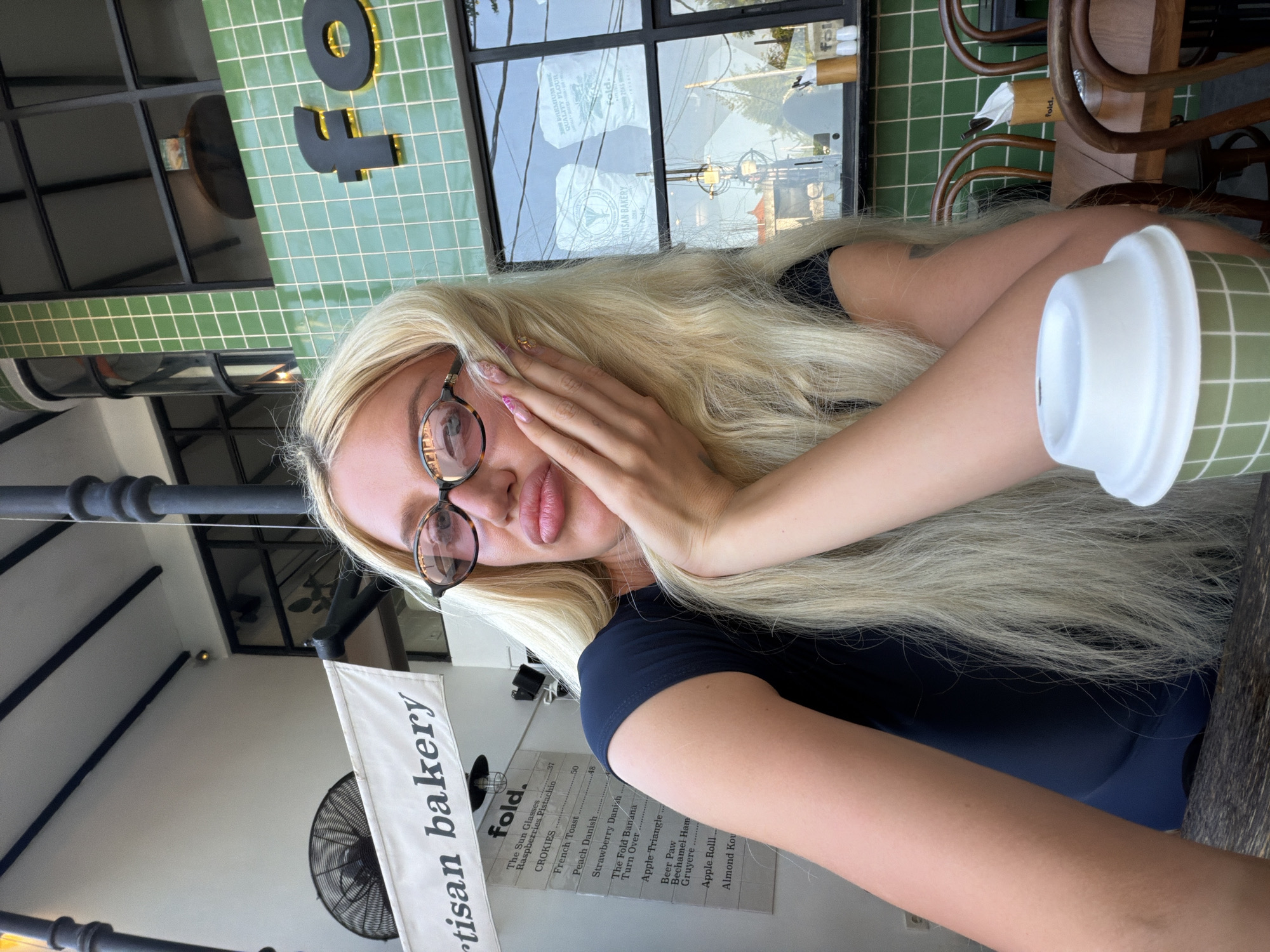 The image shows a young woman with long blonde hair and large lips, wearing glasses and a dark blue t-shirt. She is sitting at a table outside a cafe. Behind her, there is a green tiled building with the word "fold" visible in large black letters. A menu board with various food items is also visible. A coffee cup with a white lid sits on the table in front of her. The background suggests an urban environment, possibly in North Kuta, Indonesia.