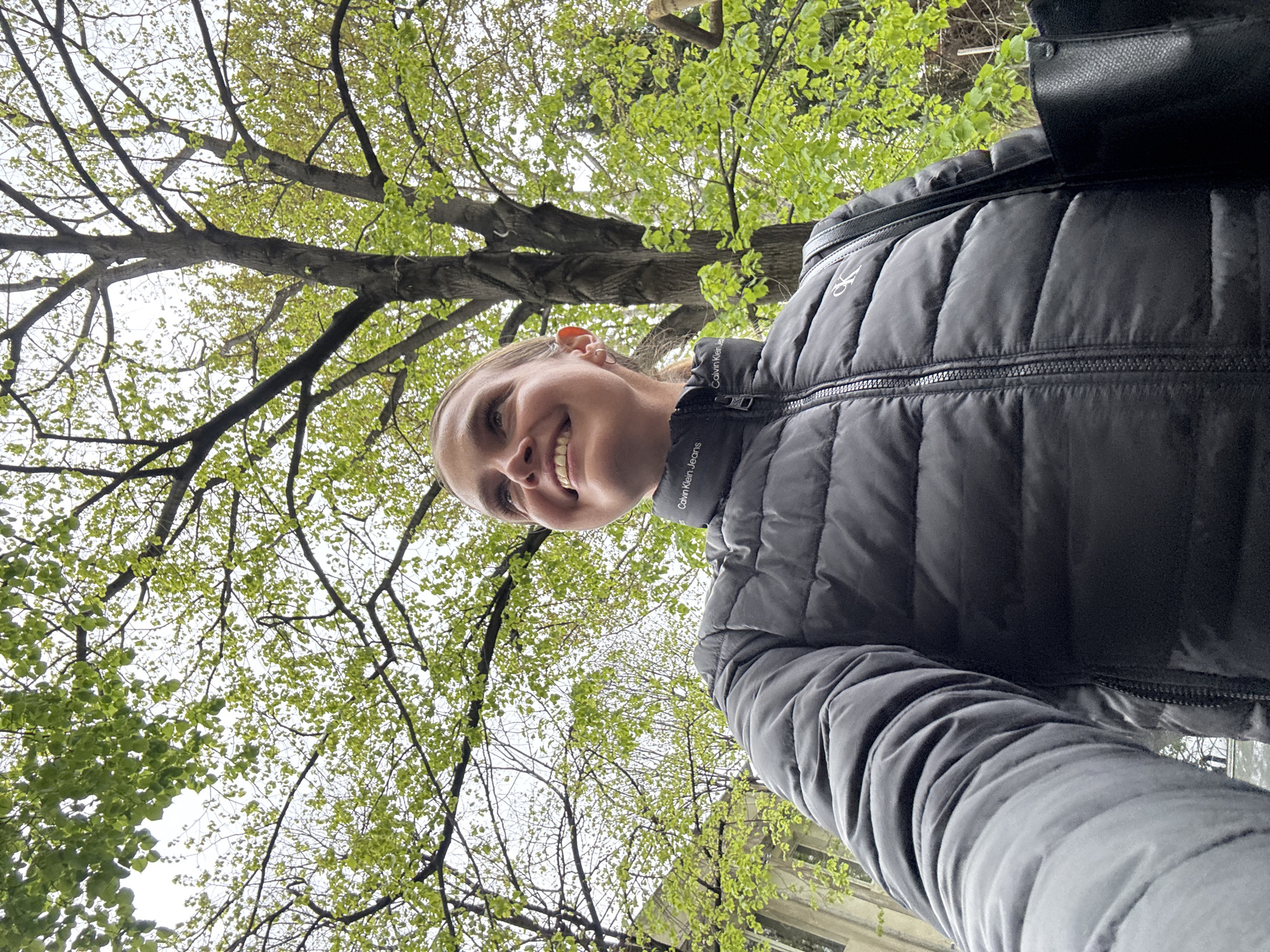This low-angle, selfie-style portrait captures a young woman outdoors in what appears to be Tbilisi, Georgia. The woman, with fair skin and light-colored hair, is smiling broadly, revealing her teeth, and looks directly towards the camera with a cheerful expression. She is wearing a dark, likely black or charcoal grey, quilted puffer jacket. The collar of her jacket features the text "Calvin Klein Jeans," and a distinct "CK" logo is visible on the strap of a black bag she carries on her right shoulder.
The background is dominated by the vibrant green canopy of a tree, its branches filled with numerous fresh, light green leaves, suggesting the season is spring. The sky above is bright and appears overcast, creating soft, diffused lighting. In the lower right portion of the background, a light-colored building or architectural structure is partially visible, indicating an urban or park setting. The overall mood conveyed by the woman's smile and the fresh greenery is one of happiness and an appreciation for the outdoors.