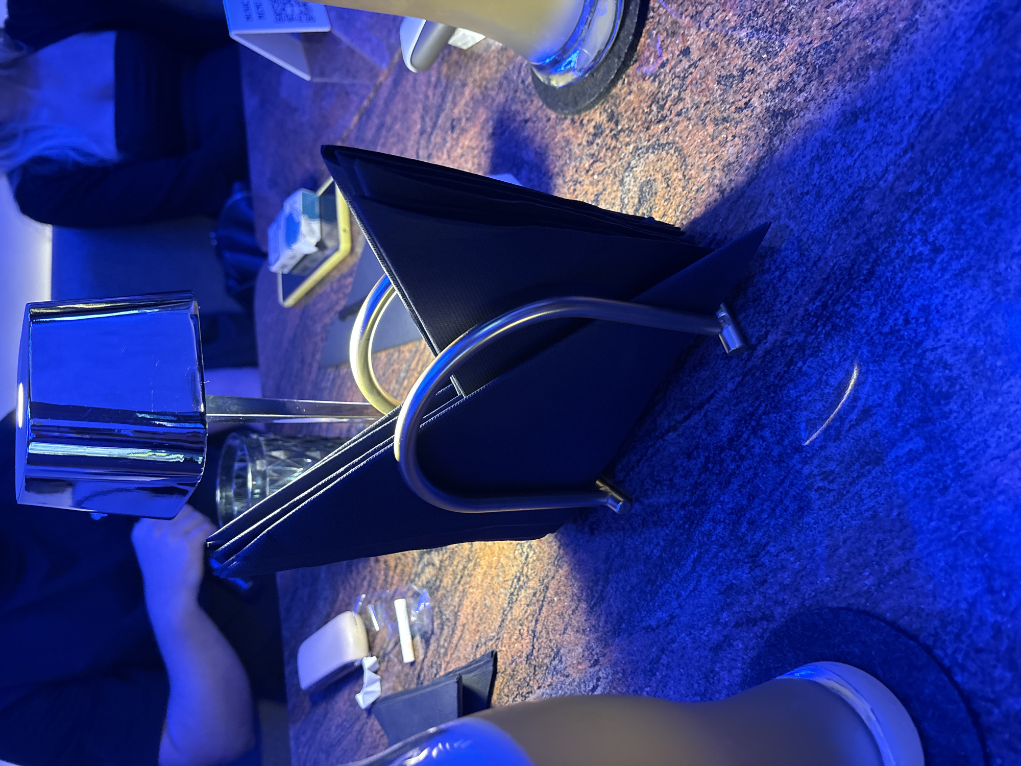 This is a close-up, medium shot of a table at night, illuminated by blue lighting. The table surface has a granite-like texture with warm and cool tones. In the foreground, a metallic napkin holder with black napkins is prominent. To the left, the base of a glass or bottle is visible. In the midground, a person wearing a black shirt is visible from the shoulders up, with their hands obscured. Another person, also wearing a black top, is partially visible in the background to the right. A yellow smartphone is on the table near a small white device and a piece of paper with a QR code. A lamp with a chrome shade is positioned behind the napkin holder. The overall impression is that of a bar or restaurant setting in Budva, Montenegro.