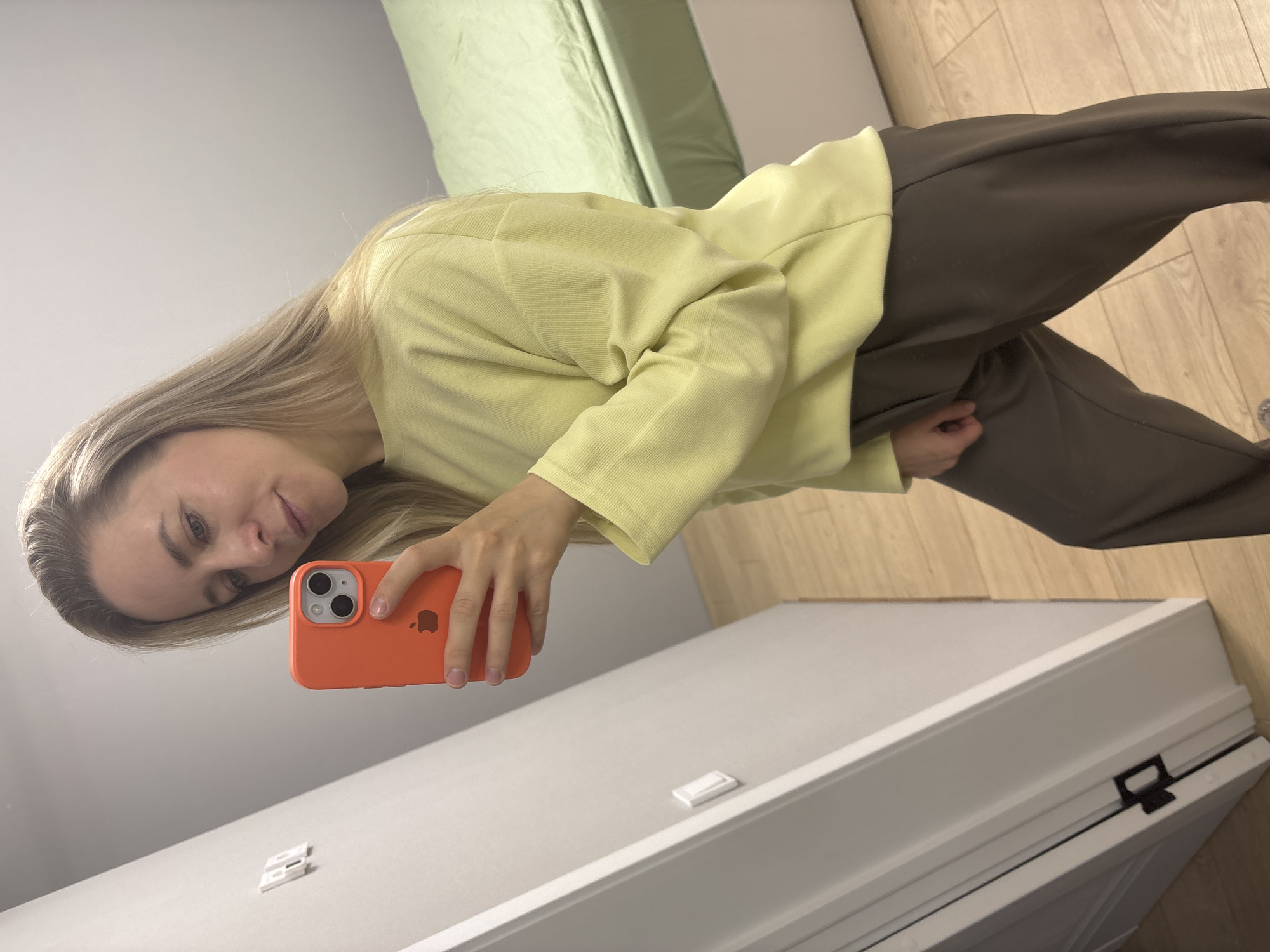 A young woman with long, straight blonde hair and light-colored eyes is captured in a mirror selfie taken indoors. She is wearing a light yellow, long-sleeved ribbed top and dark brown, loose-fitting pants. Her head is tilted slightly to the left, and she displays a gentle, subtle smile. She holds an orange-cased iPhone with both hands, positioned to take the photo. The Apple logo is visible on the back of the phone case. The background includes a plain light gray wall, a section of a light green upholstered object (possibly a headboard or sofa), and light-toned wooden flooring. In the foreground, a white dresser or cabinet with decorative molding and a small white latch or handle is visible. The setting is an indoor room in Chaikovsky, Russia.