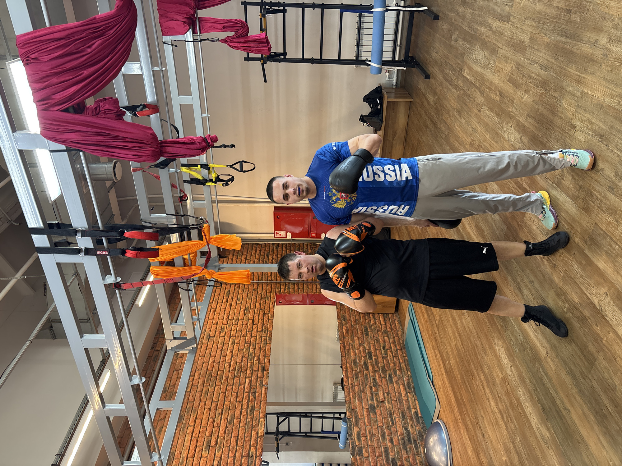 Two men wearing boxing gloves are posing for a picture in a gym setting. The man on the left is wearing a black shirt and shorts with black socks and shoes. The man on the right is wearing a blue shirt with "Russia" printed on it, gray pants, and athletic shoes. The gym features aerial yoga equipment, a brick wall, and various exercise apparatus. The gym is in Chaikovsky, Russia, as indicated by the inscription on the blue shirt.