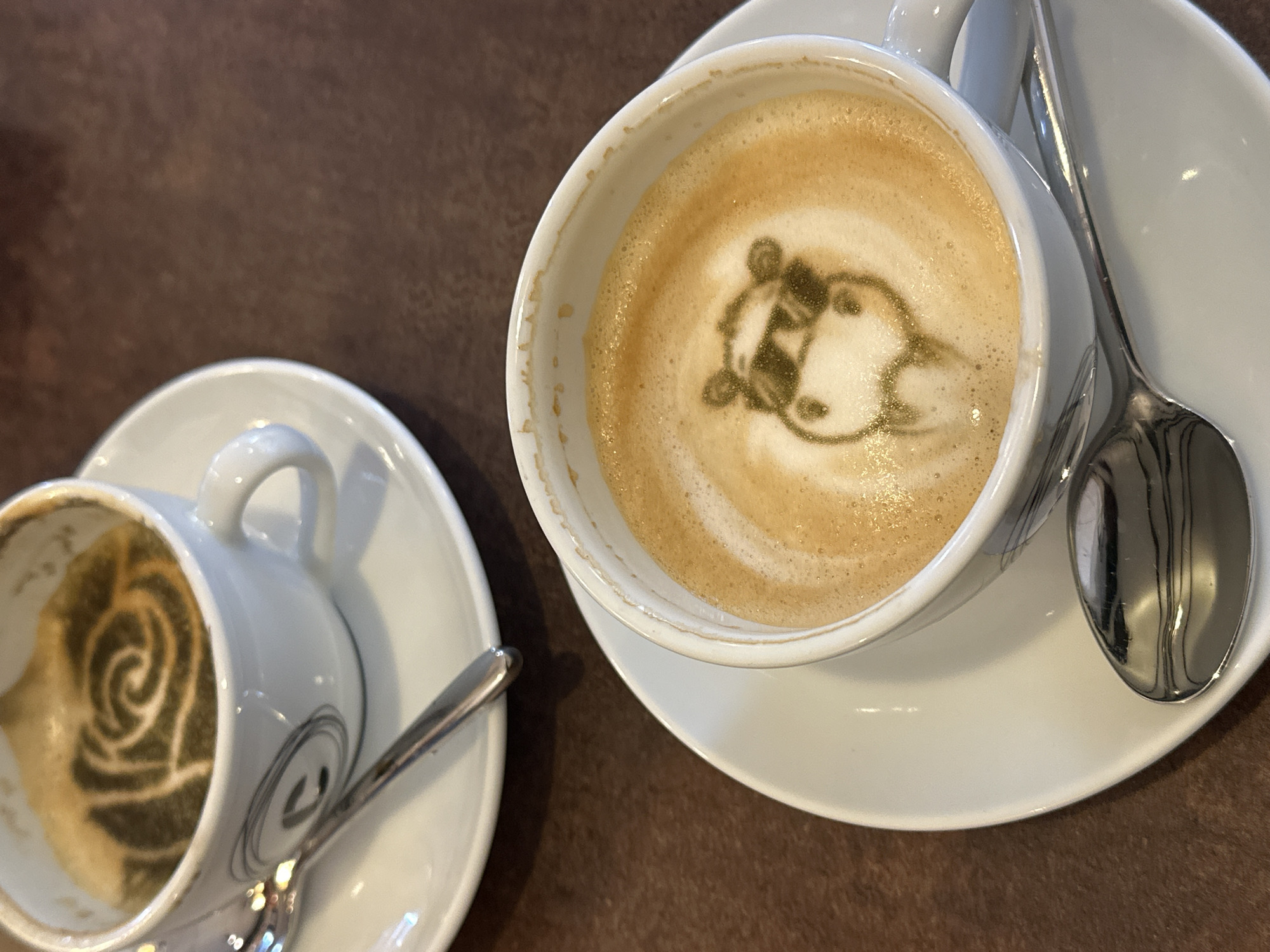The scene, located in Šušanj, Montenegro, depicts two white ceramic coffee cups with saucers and spoons placed on a dark brown tabletop. In the foreground, a white cup holds a light brown beverage with foam art depicting a stylized animal head, resembling a cow, wearing prominent dark-outlined sunglasses. This latte art is rendered in a darker green-brown substance. A silver spoon rests on the white saucer to the right of this cup. In the background, slightly out of focus, a second white ceramic cup contains a darker beverage with foam art forming a multi-layered spiral or rose pattern, also in a dark green-brown substance. This background cup exhibits a grey circular logo or design on its side. A silver spoon lies on its white saucer to the left. The uniform dark brown surface of the table suggests an indoor setting.