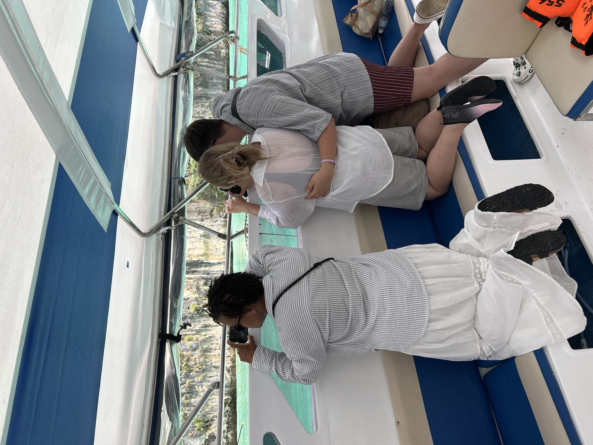 Three individuals are on a boat, looking out at a rocky coastline with turquoise water. The person on the left, a woman with dark curly hair, is wearing a grey and white striped long-sleeved shirt and a long white skirt. She is kneeling and holding a smartphone to her eye, as if taking a picture or video. In the middle, a woman with blonde hair tied back in a ponytail, wearing a sheer white shirt and grey shorts, is also kneeling. A man with short brown hair, wearing a grey striped short-sleeved shirt and red striped shorts, is standing behind her, with his arms around her waist. The boat has blue and white cushioned seating and a blue and white striped canopy overhead. In the background, steep, vegetated cliffs rise from the water. The scene appears to be a scenic tour in Ao Nang, Thailand, likely a boat trip to nearby islands or coastal areas.