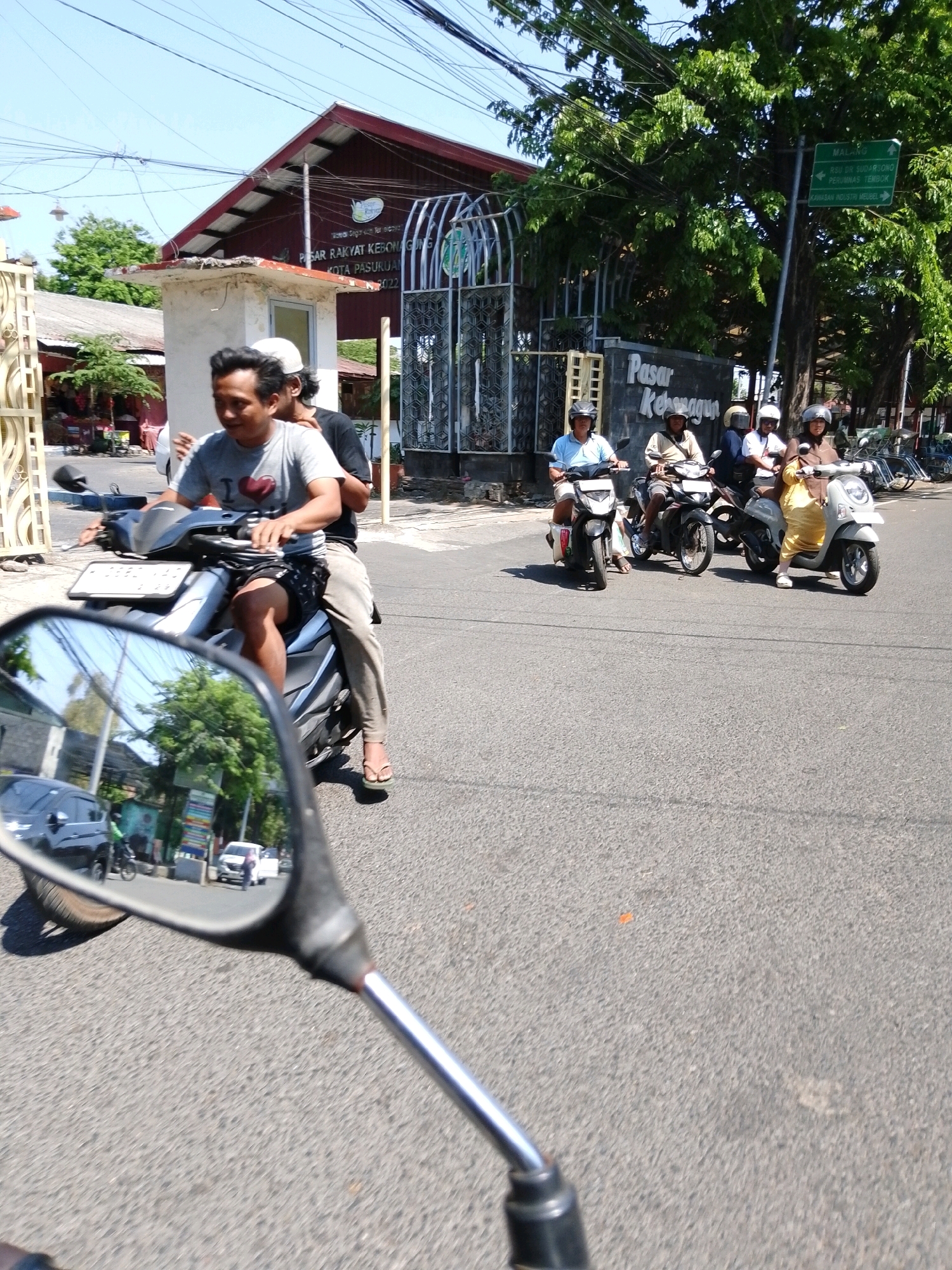 In Pasuruan, Indonesia, on a sunny day, two men are riding on a motorcycle. The rider in the front, wearing a gray t-shirt with a heart graphic and black shorts, is looking forward and smiling. Behind him, another man in a black t-shirt and light-colored pants is also on the motorcycle. The motorcycle is moving on a paved road, with other motorcycles and people visible in the background, along with buildings and trees. The scene appears to be near a market area, indicated by the "Pasar Rakyat Kebonagung" signage. The foreground shows a motorcycle's rearview mirror reflecting the road and other vehicles.