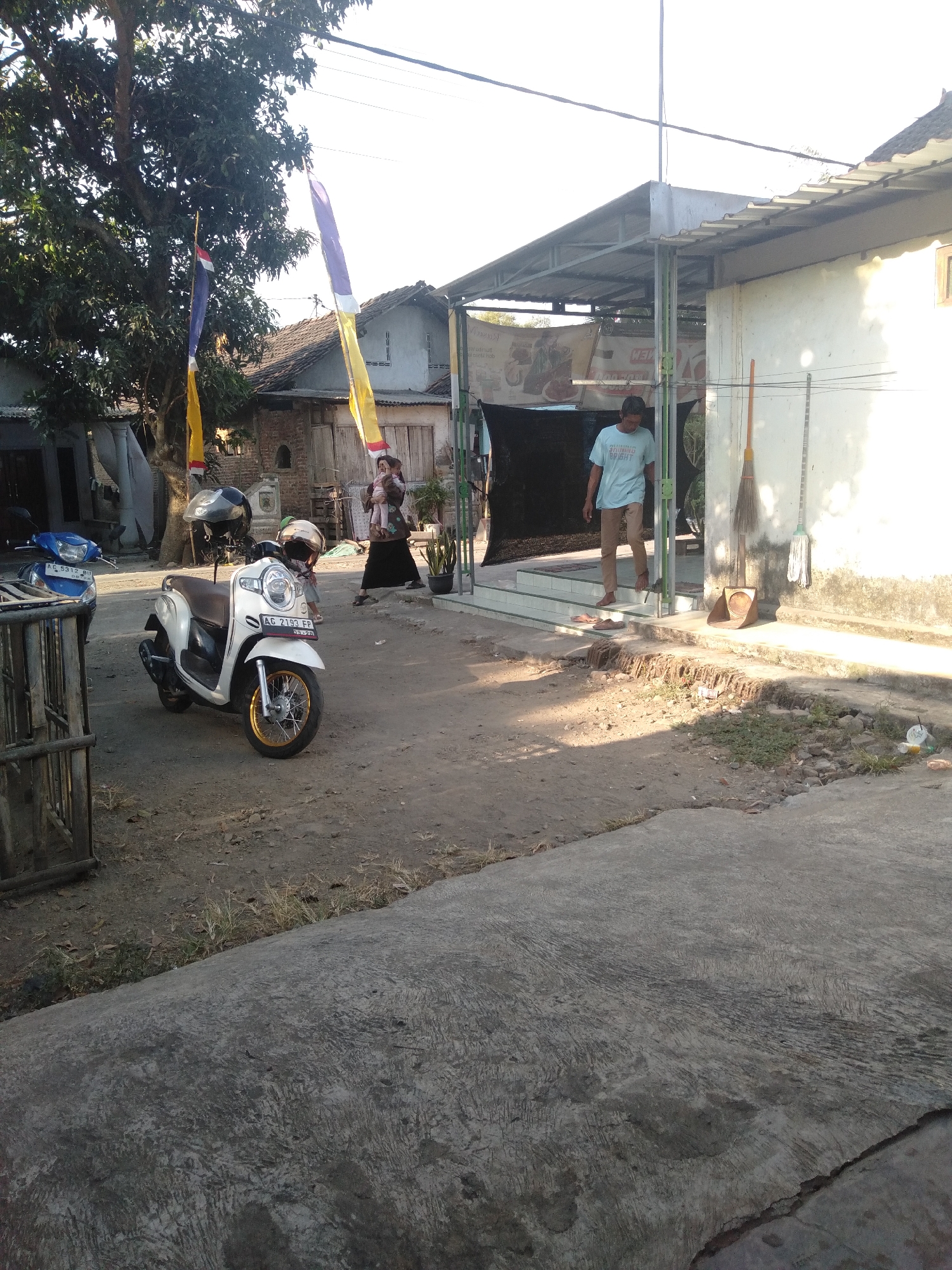 In Badas, Indonesia, a scooter, a blue motorcycle, and two individuals are visible in an outdoor setting. A white scooter with gold rims is parked on a dirt path, with a black helmet on its seat and another helmet leaning against its handlebars. To the left, a blue motorcycle is partially visible behind a wooden fence. A woman in a headscarf and dark clothing is carrying a child. A man in a light blue t-shirt and tan pants is walking down steps. Two brooms and a dustpan are propped against a white building with a corrugated metal roof. In the background, trees and traditional Indonesian houses with tiled roofs are present. The sky is bright and clear.