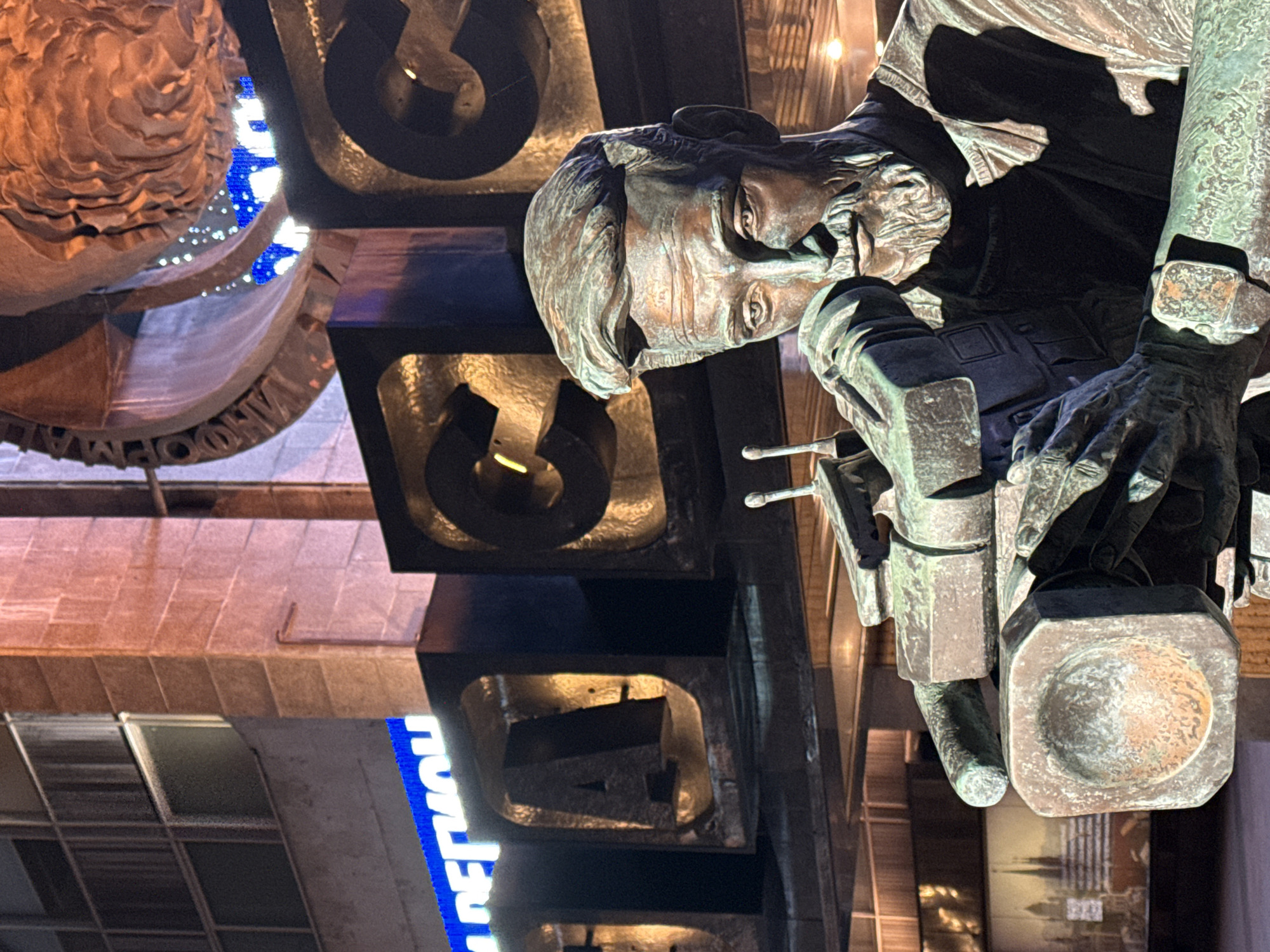 The image features a close-up of the Monument to the Conquerors of Space in Moscow, Russia. The sculpture depicts a stylized figure of Konstantin Tsiolkovsky, a Russian and Soviet rocket scientist and astronautics pioneer, standing next to a large abstract structure with Cyrillic letters spelling out "To the Conquerors of Space." Tsiolkovsky is looking down and holding a rectangular object. The scene is captured at night, with lighting illuminating the monument. Background details include buildings and a blue sign with Cyrillic lettering.