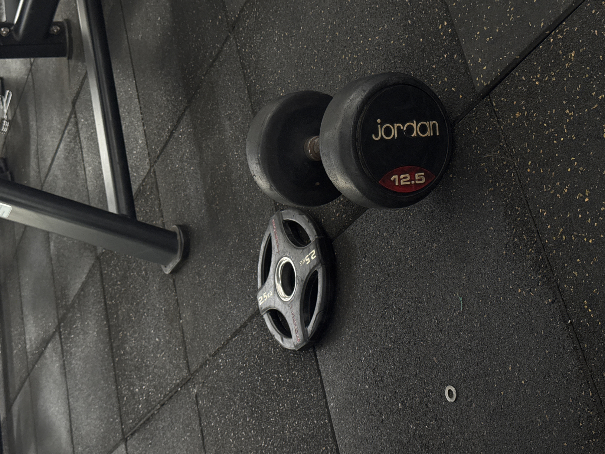 An overhead shot captures a gym floor featuring a black dumbbell and a weight plate. The dumbbell, branded "Jordan," is marked with "12.5" on its end. To its left, a black weight plate labeled "2.5 KG" is placed on the textured black floor, which is composed of interlocking rubber tiles. In the background, the legs of gym equipment are visible. The scene is lit by overhead lighting, casting faint shadows. There are no people present.