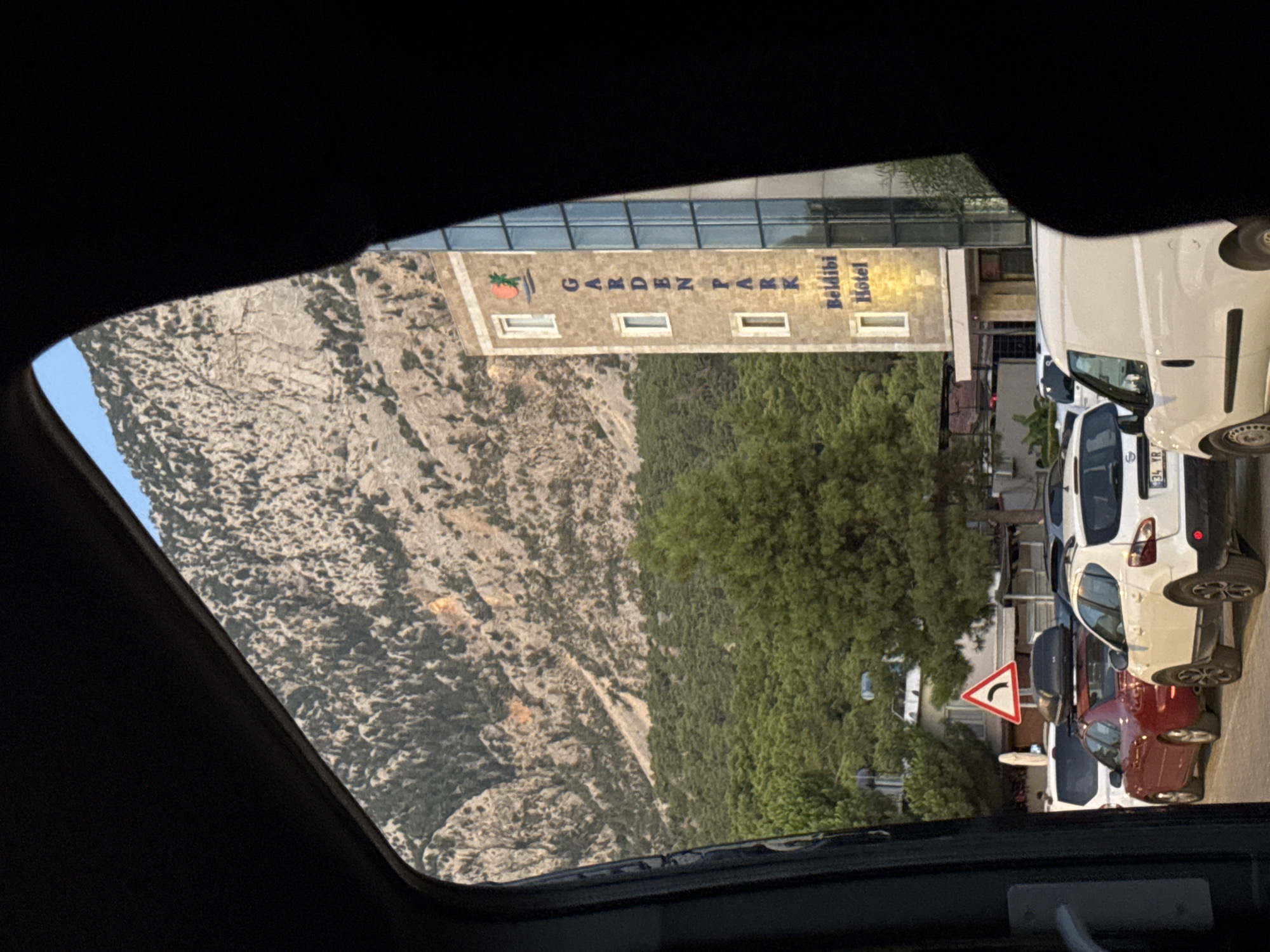 The image shows a view from inside a vehicle looking out towards a street scene in Kemer, Turkey. A large, rocky mountain dominates the background, covered with sparse vegetation. In the middle ground, a multi-story building with "GARDEN PARK Beldibi Hotel" written vertically on its facade stands out. The building has windows and a glass-paneled section. In front of the hotel, a dense forest of green trees is visible. The foreground of the image features several vehicles parked and moving on a road. These include a white van, a white SUV with a license plate partially visible, a red sedan, and another white vehicle. A triangular traffic sign with a curved arrow indicating a turn is visible. The sky above is clear and blue. The overall scene suggests a daytime setting in a town nestled between mountains and the coast.