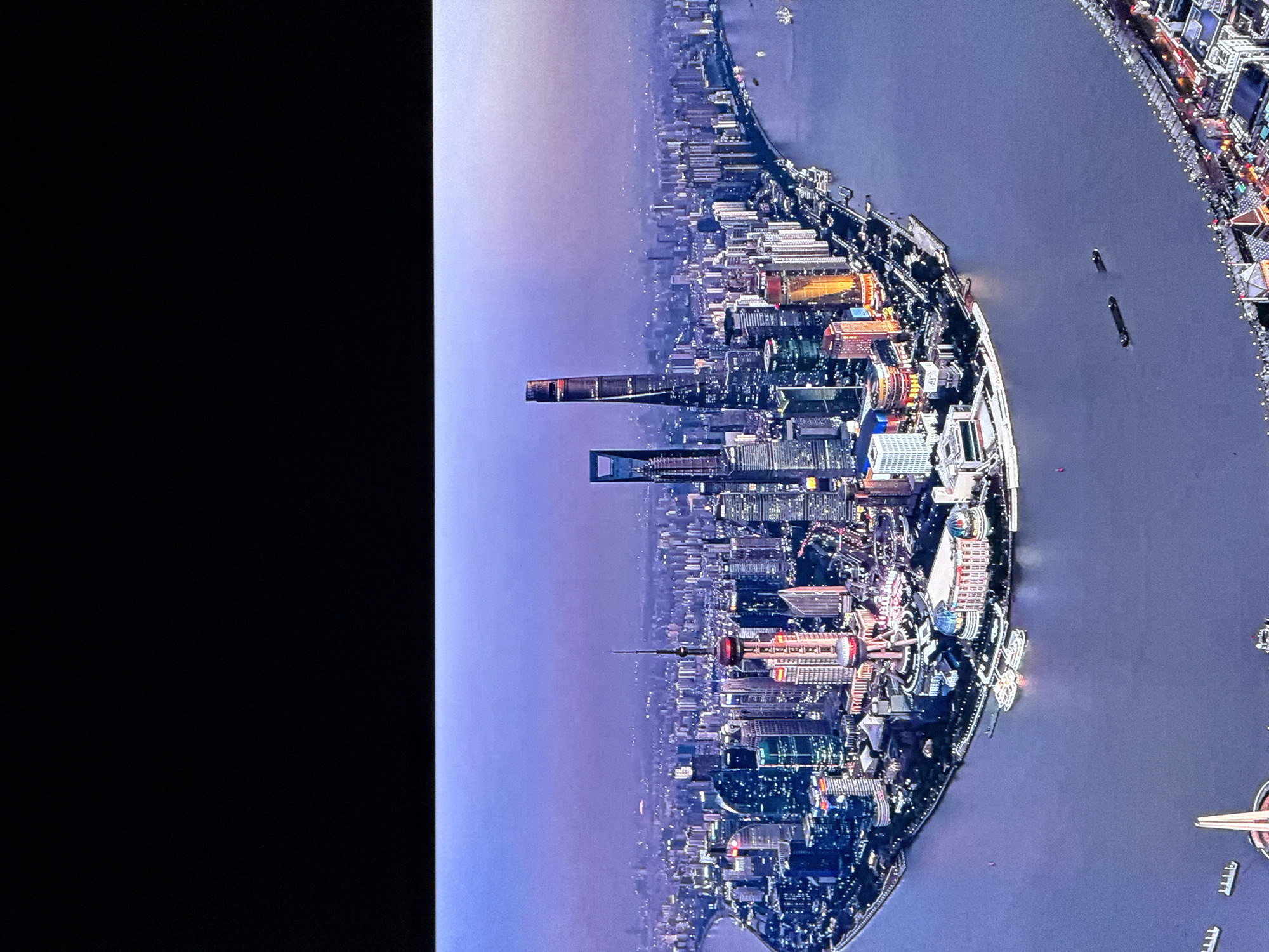The image is an aerial view of a sprawling city skyline at dusk, featuring numerous skyscrapers and a wide river in the foreground. The buildings are illuminated with a mix of white, blue, and orange lights, creating a vibrant urban landscape. The Oriental Pearl Tower, a distinctive landmark with its spherical structures, is prominently visible among the skyscrapers. Several boats are present on the river, some docked and others in transit. The sky is a hazy purple, suggesting either pollution or the twilight hour. The overall impression is of a busy and modern metropolis at night. The scene depicted is Shanghai, China, not Saint Petersburg, Russia.