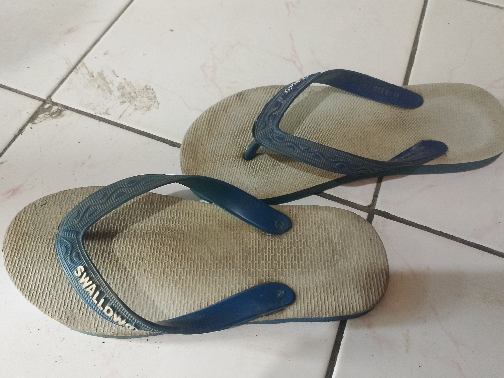 A pair of used, blue and tan flip-flops are lying on a tiled floor. The flip-flops are placed parallel to each other, with the left flip-flop slightly in front of the right. The left flip-flop has the word "SWALLOW" embossed on its strap. The right flip-flop has "SIZE: 11" embossed on its strap. The floor is made of white tiles with dark grout lines. There are some dirt smudges on the tiles. The image was taken in Mukapayung, Indonesia.