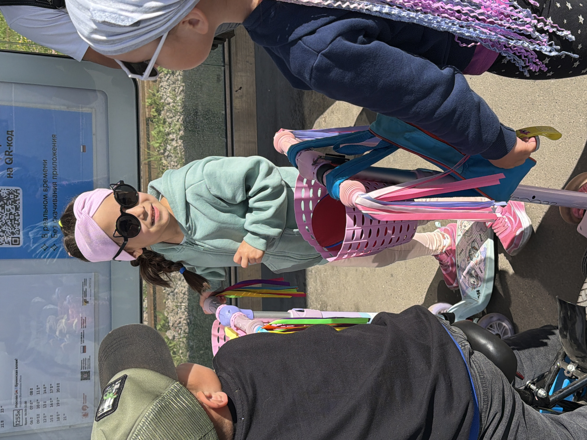 In Shcherbinka, Russia, three children are gathered around scooters on a paved surface, likely a sidewalk or pathway. The child in the center, wearing a mint green hooded sweatshirt, a light pink headband, and black cat-ear sunglasses, is holding onto the handlebars of a light blue and pink scooter adorned with colorful streamers. To her left, a child wearing a black long-sleeved shirt and a green baseball cap is facing away from the camera, positioned next to a scooter. To her right, a child in a navy blue hooded sweatshirt and a light gray headscarf is holding onto the handlebars of another scooter. The background features a large glass panel with text and a QR code, possibly a public transport information display, and a glimpse of green foliage. The lighting suggests it is daytime.