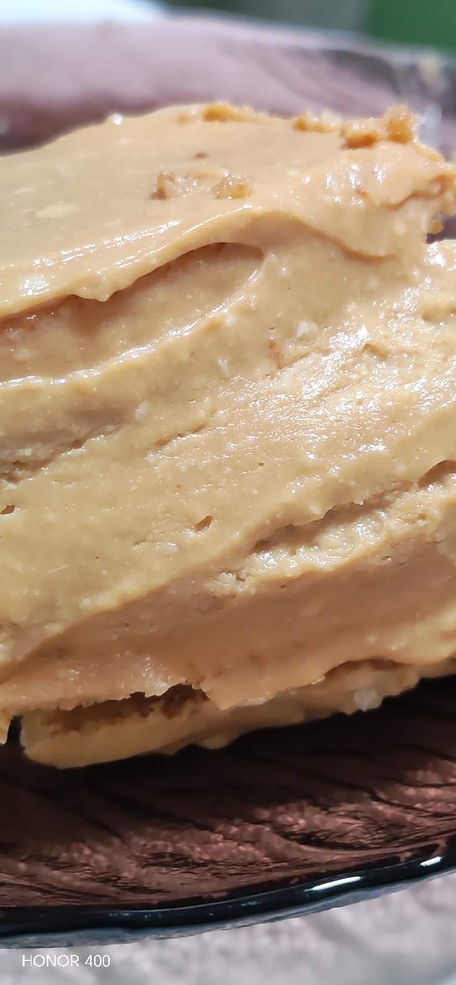 The image shows a close-up of a layered dessert, possibly a cake or a layered bar, with a smooth, light brown topping. The layers appear to be uneven, with some visible gaps. The dessert rests on a dark-colored plate. There is no indication of the location being Noviy, Russia, or any other specific place. The image contains no people, actions, or interactions.