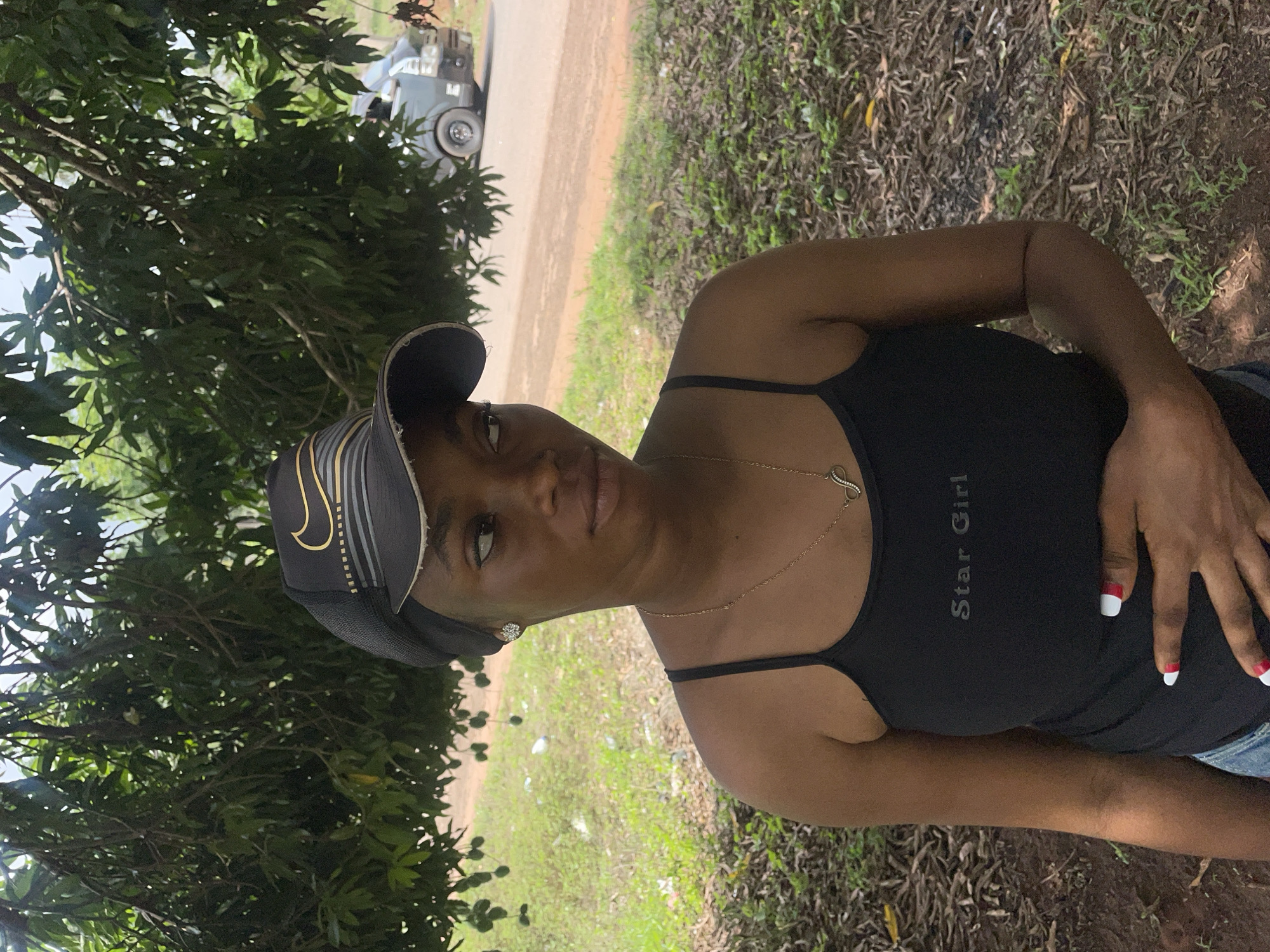 A woman stands in an outdoor setting. She wears a black tank top with "Star Girl" printed on it, a gold necklace, and a black cap. The surrounding environment includes trees, grass, and brown foliage on the ground. A vehicle is partially visible in the background. This is in Ogbe, Nigeria.