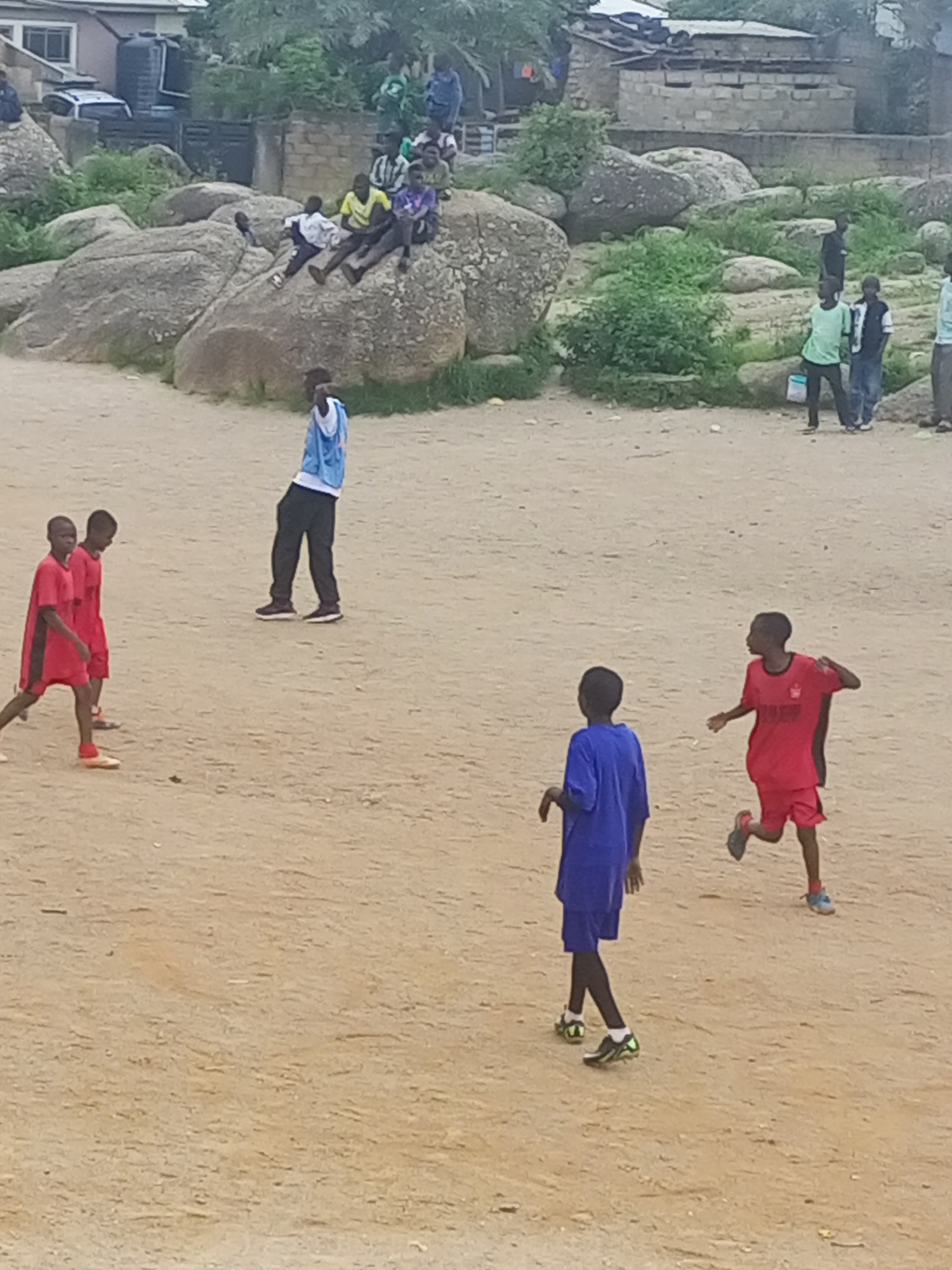 This image captures a street scene in Jos, Nigeria, where several young boys are engaged in a game of football on a dusty, unpaved area. The background features rocky outcrops, some scattered vegetation, and a few simple structures, suggesting a semi-urban or fringe-of-city setting.
In the foreground, two boys in red football kits are positioned on the left, seemingly involved in play. One of them, further back, appears to be dribbling the ball. To their right, a boy in a blue jersey and black pants stands facing away from the camera. In the mid-ground, a boy in a white and blue striped shirt and black pants sits on a large rock, facing the game. Further to the right, a boy in a red football kit is in motion, looking to his left as he runs. Behind him, another boy in a blue outfit is also running.
In the background, several other individuals are present, some standing and observing the game, others seemingly interacting or walking. A white vehicle is parked near a building in the upper left of the image. The overall atmosphere suggests a casual, informal gathering for sport and recreation.