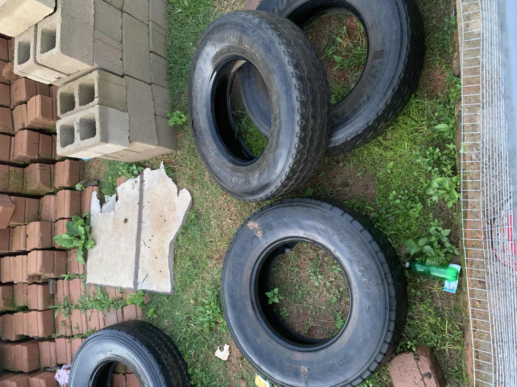 In a grassy yard, four tires are stacked on top of each other. A pile of red bricks and concrete blocks are visible in the background, along with a piece of broken concrete. A green plastic bottle and a white object are on the ground in front of the tires. A wire fence runs along the bottom of the image.