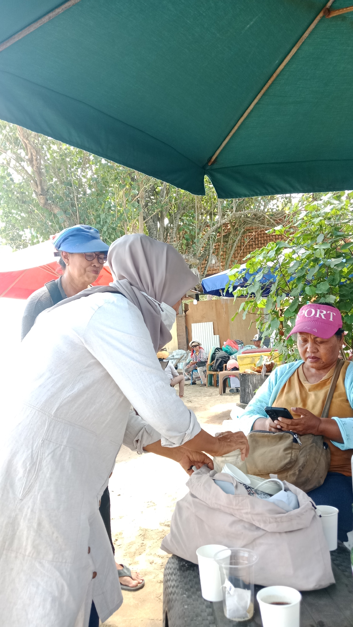 In Kuta, Indonesia, under a large green umbrella, two women are engaged in an interaction. The woman on the left, wearing a blue cap and glasses, appears to be looking at the woman in front of her. The woman in front, wearing a grey hijab and a light-colored garment, is reaching into a large light-colored bag, holding some white items, possibly cups or utensils. To the right, another woman in a yellow shirt and a light blue cardigan, wearing a pink visor with "PORT" written on it, is looking down at her phone and holding a brown bag. In the background, other people are seated, and there are trees and structures visible. The ground appears to be sandy.