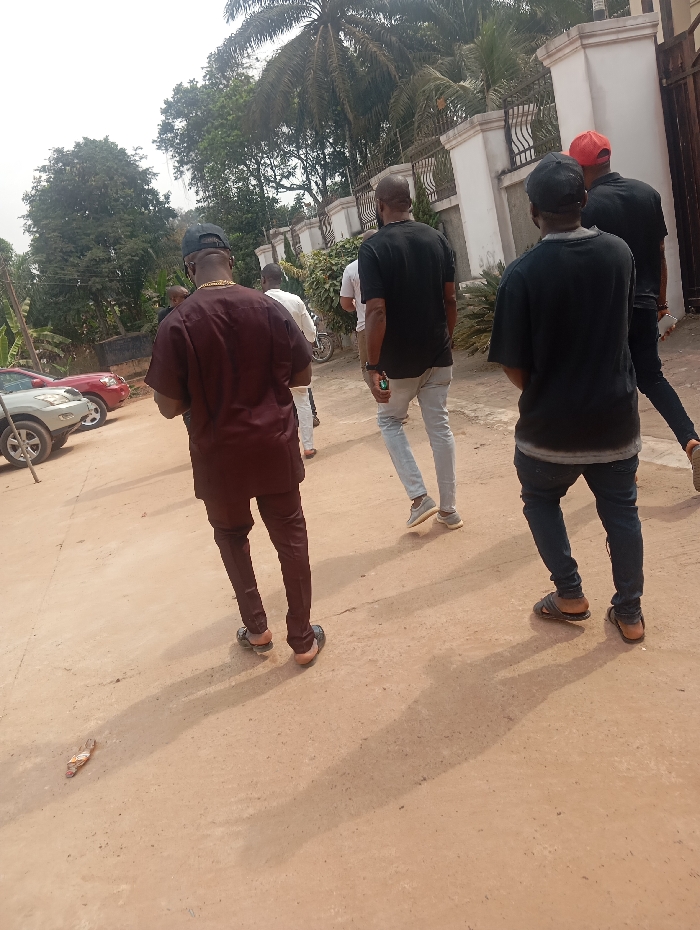 The scene depicts a group of approximately five to six adult males walking on an unpaved, reddish-brown dirt path in Okpatu-Ukehe, Nigeria. The men are largely viewed from behind, moving generally towards the right side of the frame. In the immediate foreground, a male wears a dark maroon traditional top and trousers with dark sandals, a black cap, and a visible gold-colored chain. To his right and slightly behind are other men: one in a white shirt and light trousers, another in a black t-shirt and light blue jeans with sneakers, another in a black t-shirt, dark trousers, and a black cap with dark sandals, and a man furthest right wearing a red cap, black shirt, and dark trousers. A single detached light-colored flip-flop lies on the ground in the lower left.
The environment features lush green vegetation, including tall trees and a prominent palm tree, behind a white building with a decorative black wrought-iron fence and gate structure. On the far left, two parked vehicles are partially visible: a silver SUV and a red sedan. Long shadows are cast on the path from the subjects, indicating direct sunlight. No direct interactions between the individuals are discernible.