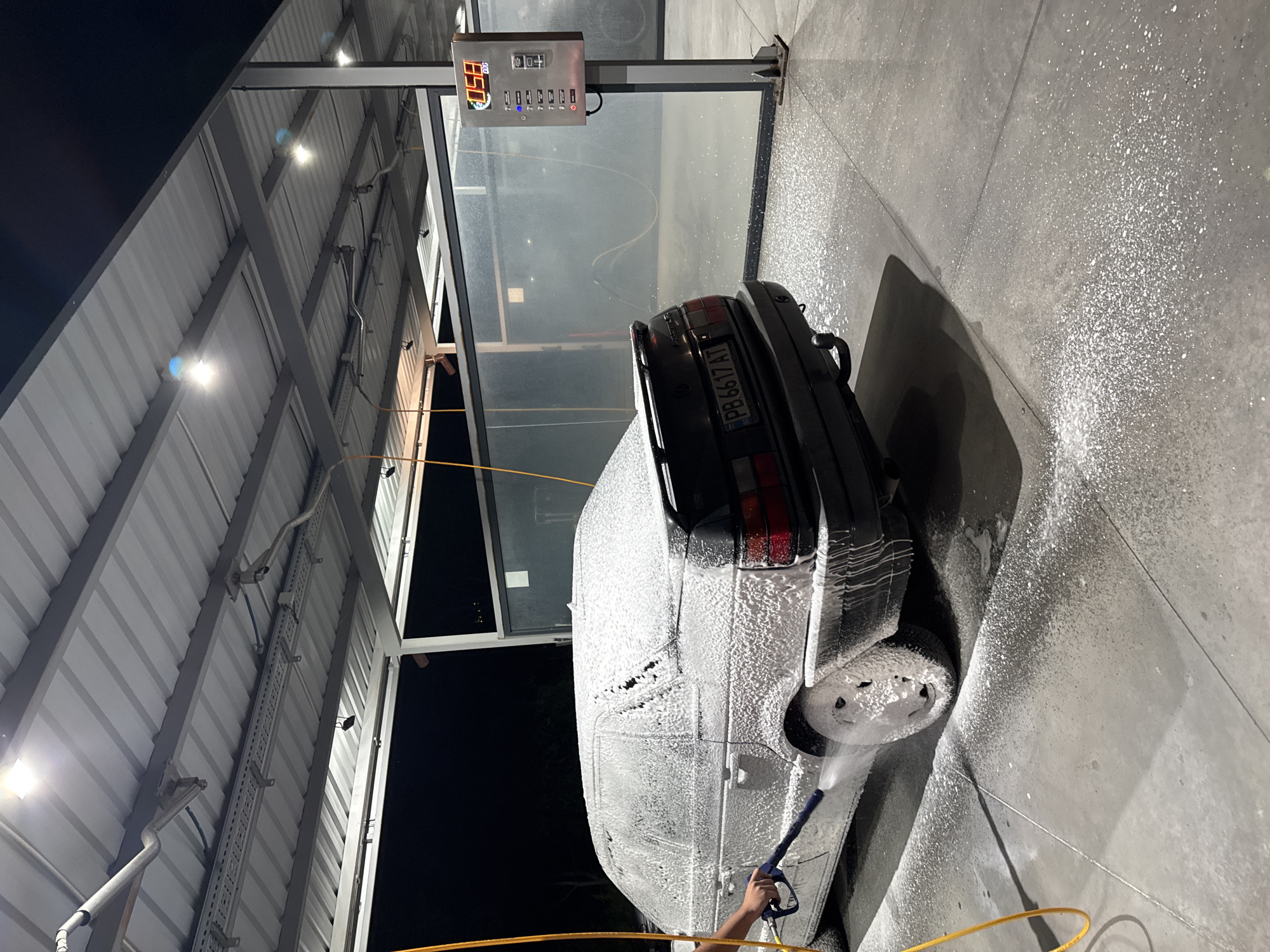 The image shows a car being washed at a self-service car wash. The car is covered in soap foam. A person is using a pressure washer to clean the car. The car's license plate reads "PB 6617 AT", which is a Bulgarian license plate. The location appears to be in Plovdiv, Bulgaria. The car wash has a metal frame with a roof and a timer/control panel. The ground is wet with water and soap. It is nighttime, and the area is lit by artificial lights.