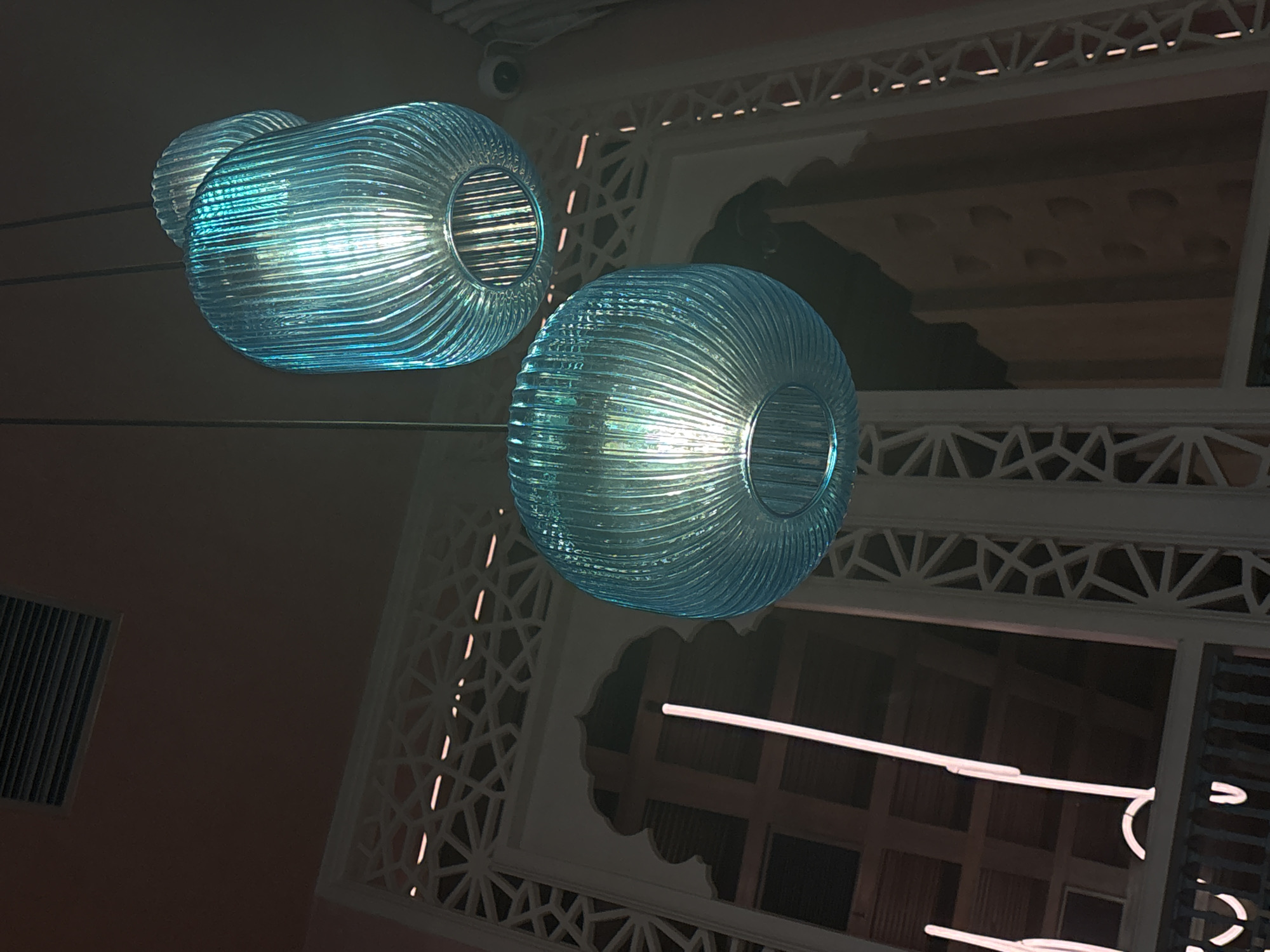 This image captures an interior architectural scene, focusing on two prominent, illuminated light fixtures. The lights are cylindrical or donut-shaped, crafted from translucent, ribbed blue-green glass that emits a bright, cool light. They appear to be suspended by wires against a dark background, with their glow being the primary light source in the immediate foreground.
The background reveals intricate white architectural elements featuring geometric patterns, possibly resembling traditional Islamic or Southeast Asian motifs. These patterns form decorative frames around what appear to be openings or windows. One opening in the mid-right exhibits a scalloped, arched shape. In the upper center, a small, black surveillance camera is visible. To the lower left, a dark rectangular vent is mounted on a wall. Towards the lower right, sections of neon-like strip lighting can be seen, adding to the artificial illumination of the space.
The overall setting suggests a modern establishment, such as a restaurant, hotel, or lounge, in North Kuta, Indonesia, incorporating local design influences. The lack of natural light and the prominence of artificial illumination indicate that the photograph was taken in the evening or at night, or within an indoor space entirely reliant on artificial light. There are no people present, and no discernible activity is taking place. No visible text is present in the image.