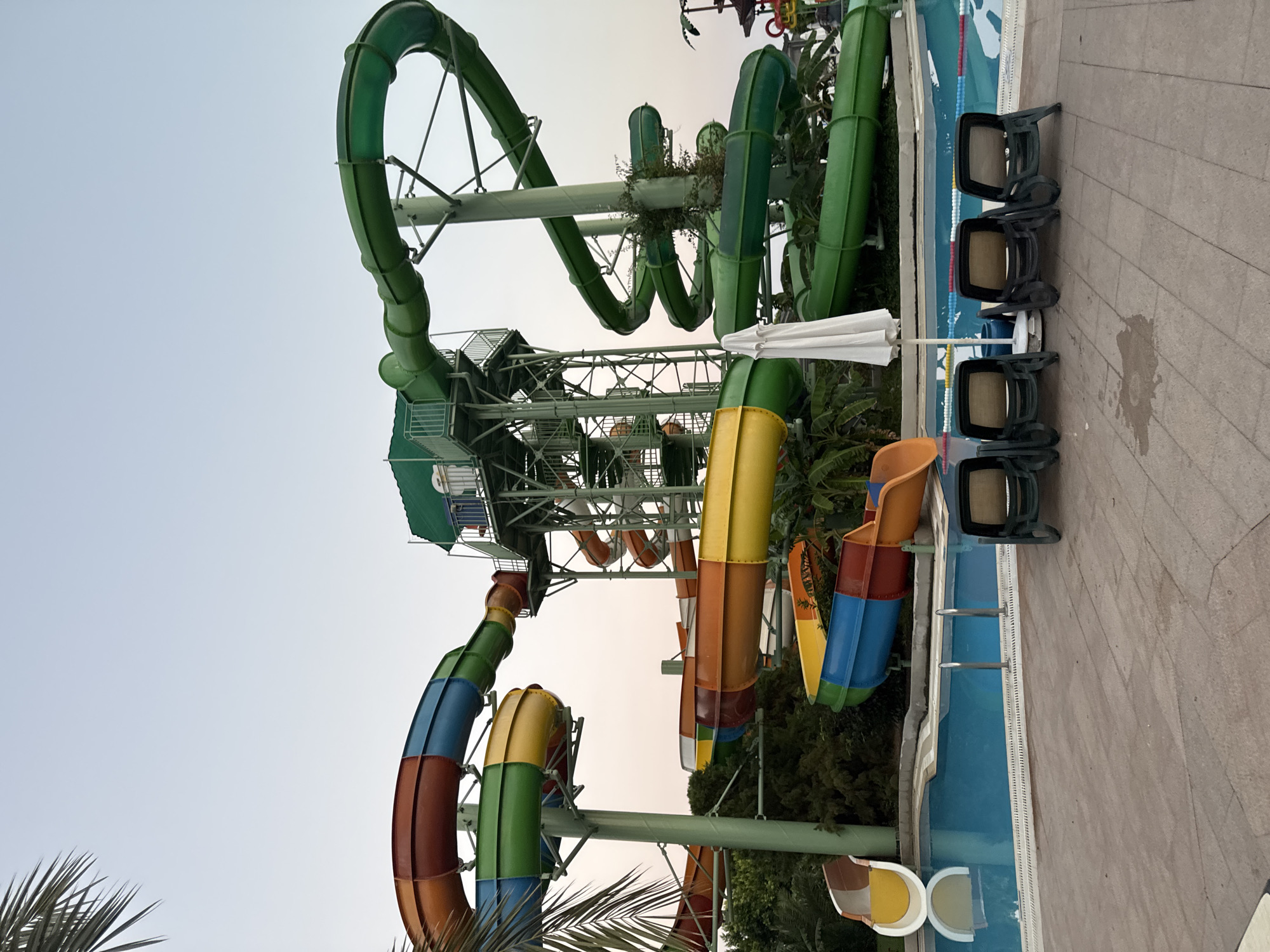 The image shows a water park with multiple colorful water slides in Aksu, Turkey. The slides are made of cylindrical sections and come in various colors including green, orange, blue, and yellow. They are supported by metal structures and wind around a central tower. A swimming pool with blue water is visible in the foreground, with lounge chairs arranged around it. Palm trees and lush greenery surround the water park. The sky is overcast, suggesting either early morning or late evening. There are no people visible in the image.