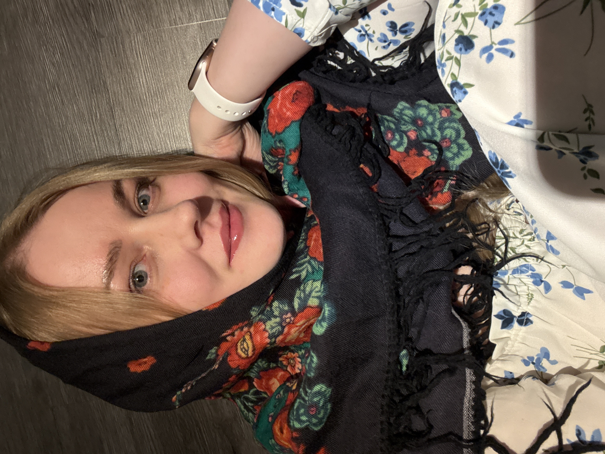 A blonde-haired woman with light skin is photographed from an unusual, slightly tilted perspective. She is wearing a white dress with blue floral patterns on the upper right side of the image. The woman has a black scarf with a floral pattern primarily composed of red and green, covering the lower left side of her face and head. The scarf has a fringed edge. She also wears a white smartwatch on her left wrist. The background appears to be a dark, wooden surface. The location and city information requested is not discernible from the image.