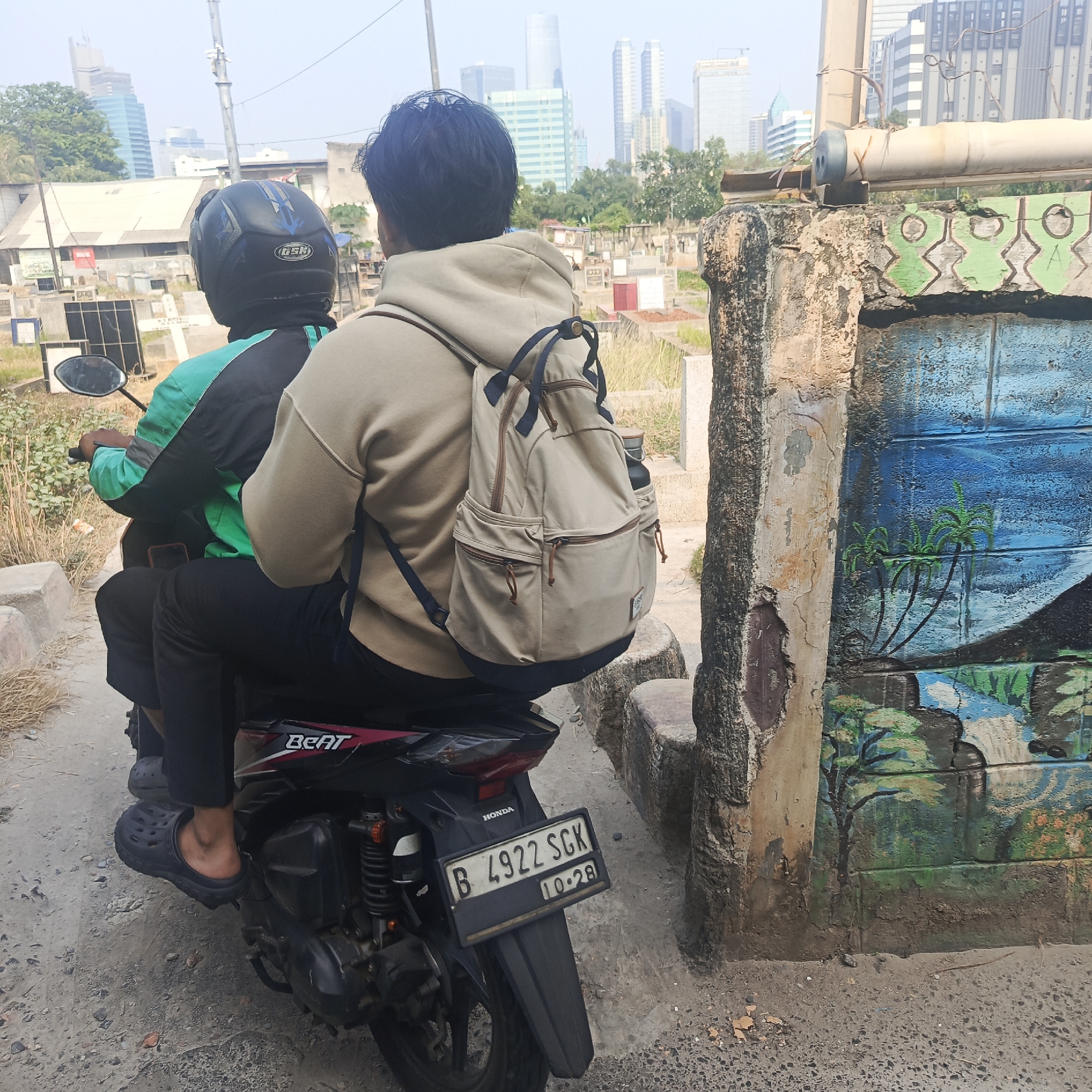 Two individuals are riding on a motorcycle in an urban environment, possibly South Jakarta, Indonesia. The rider in front wears a black helmet with blue accents and a green and black jacket. The passenger behind wears a tan hooded sweatshirt and carries a tan backpack. The motorcycle is black with red accents and a license plate reading "B 4922 SGK" with "10-28" below it. The background features a mixture of modern skyscrapers and older, low-rise buildings, suggesting a developing urban area. A concrete wall with a colorful mural depicting palm trees and water is visible on the right side of the image. The ground is unpaved and appears to be a dirt or gravel path.