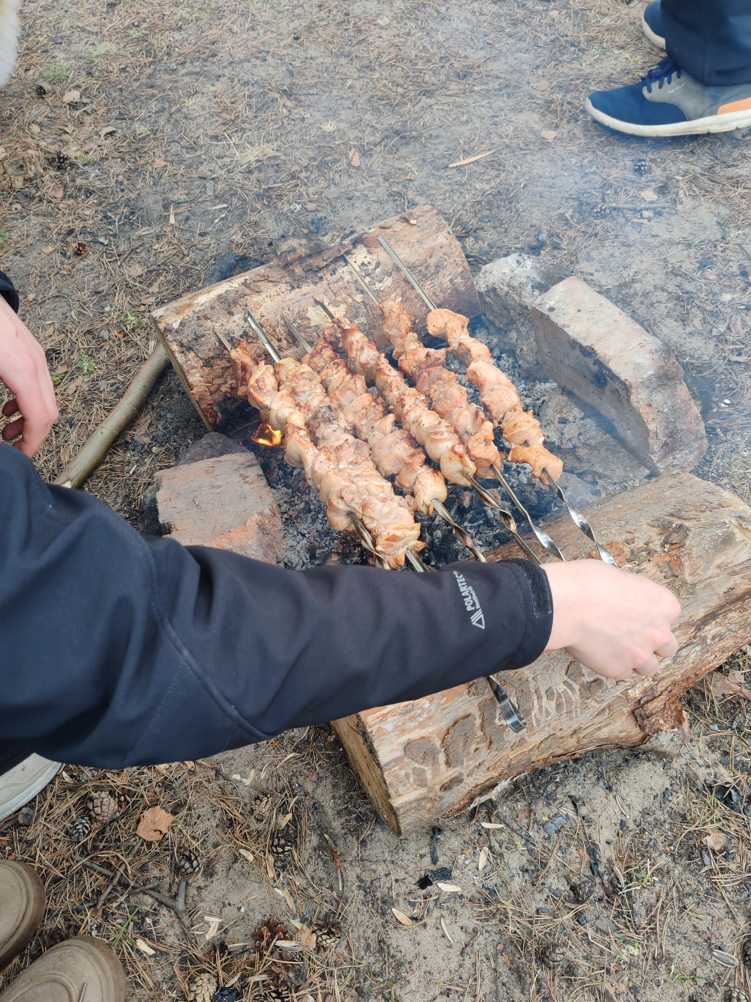 The image depicts an outdoor cooking scene, likely in a wooded area near Bolshaya Izhora, Russia. Several metal skewers laden with meat are arranged over a makeshift fire pit constructed from logs and bricks. A person, visible by their arm and hand clad in a black jacket with a PolarTec logo, is tending to the skewers. The fire is burning, and some smoke is visible. The ground is covered with pine needles and other organic debris. A pair of feet in blue and grey athletic shoes are visible in the upper right of the image, indicating another person is present. In the bottom left, a pair of beige shoes and a portion of the person wearing them can be seen.