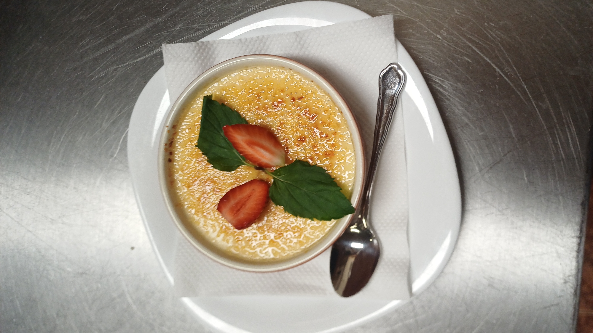 A close-up, overhead shot shows a crème brûlée dessert in a ramekin, placed on a white napkin and a larger white plate. The dessert has a caramelized sugar crust and is garnished with two halved strawberries and two green mint leaves. A silver spoon rests on the plate to the left of the ramekin. The setting appears to be a metallic surface with visible scratches. The location is Unknown, Unknown.